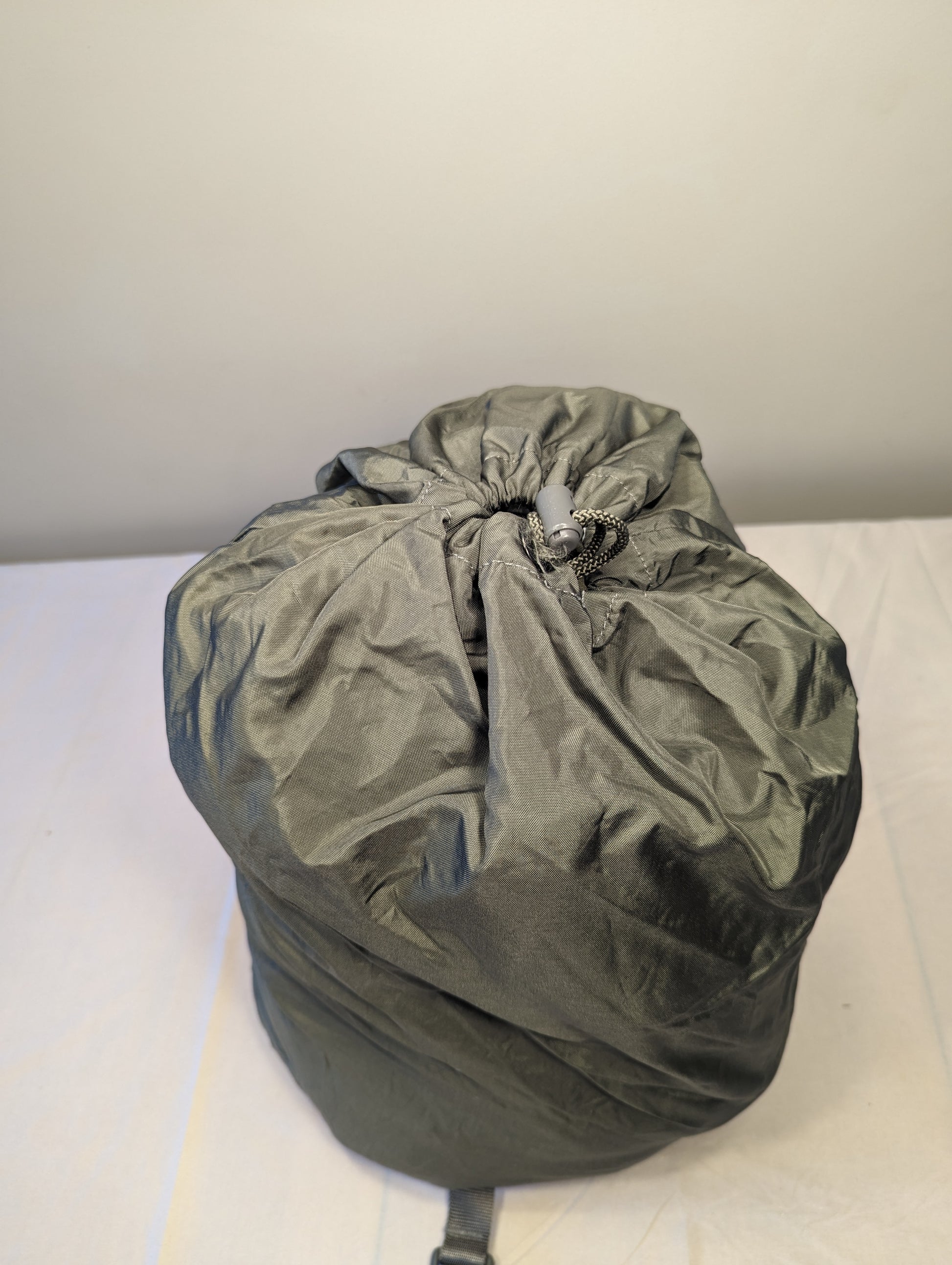 A large, tightly compressed U.S. Military Surplus Compression Stuff Sack in olive green, standing upright, featuring a cinched drawstring closure and a wrinkled nylon texture.