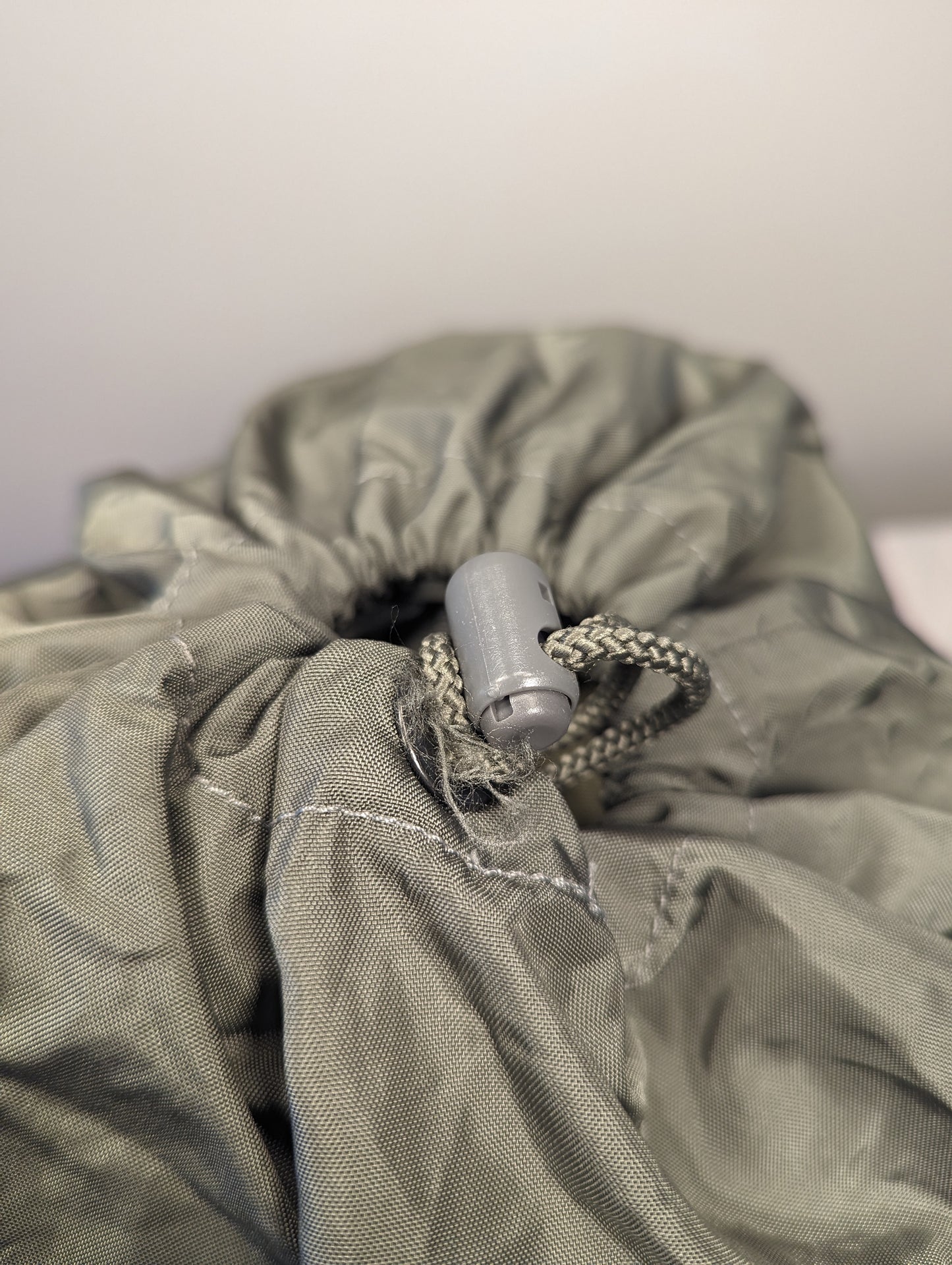 A close-up view of the drawstring closure of a U.S. Military Surplus Compression Stuff Sack in olive green, highlighting the wrinkled nylon fabric, heavy stitching, and plastic cord lock.