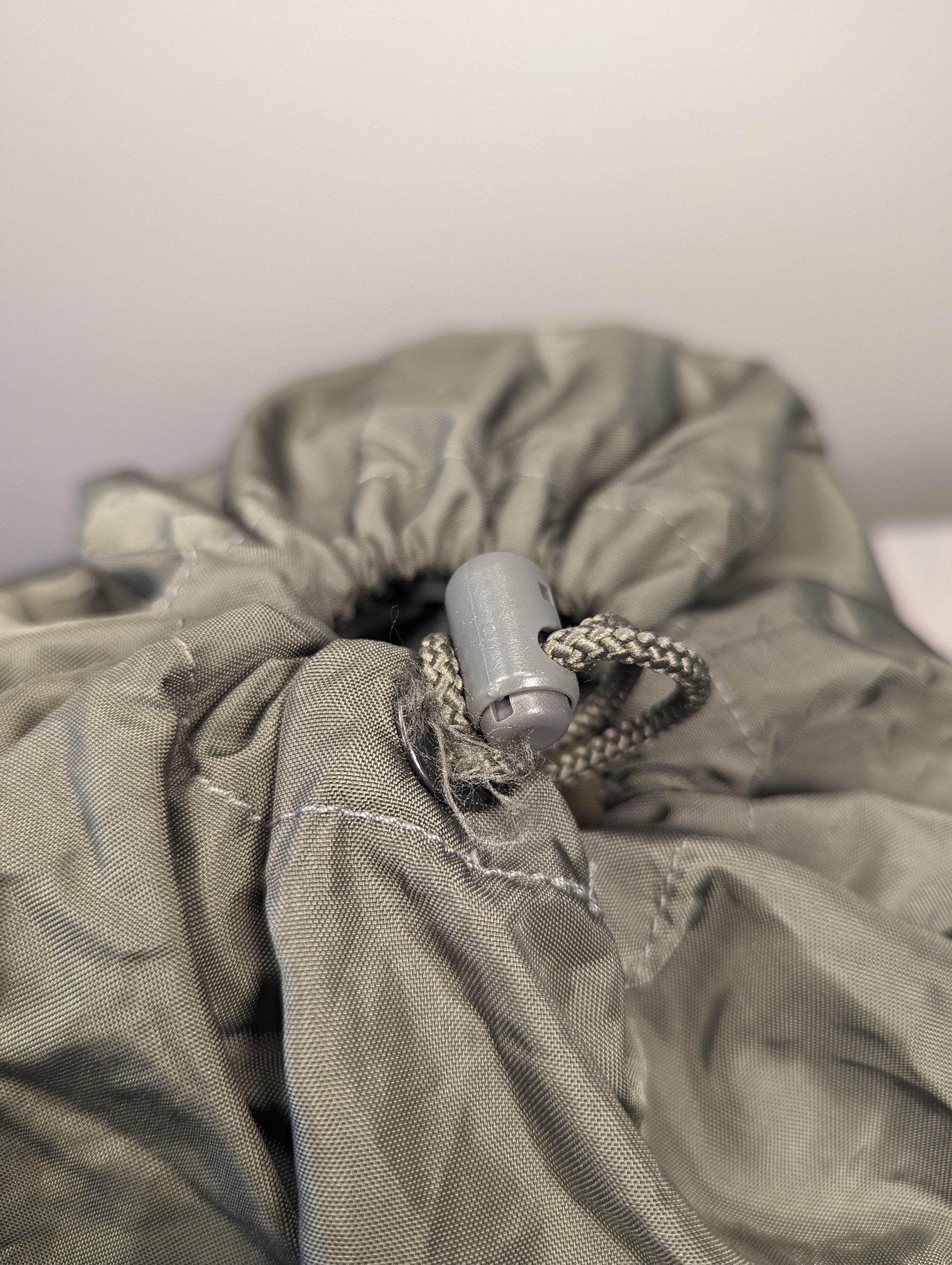 A close-up view of the drawstring closure of a U.S. Military Surplus Compression Stuff Sack in olive green, highlighting the wrinkled nylon fabric, heavy stitching, and plastic cord lock.