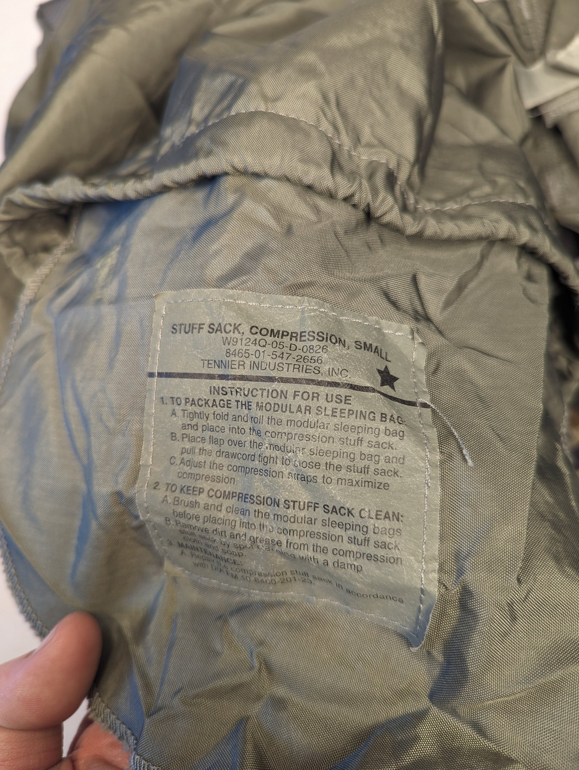A close-up photograph of the official U.S. Military issue label sewn into the olive green Compression Stuff Sack, displaying the product name, detailed packing instructions, and washing instructions.