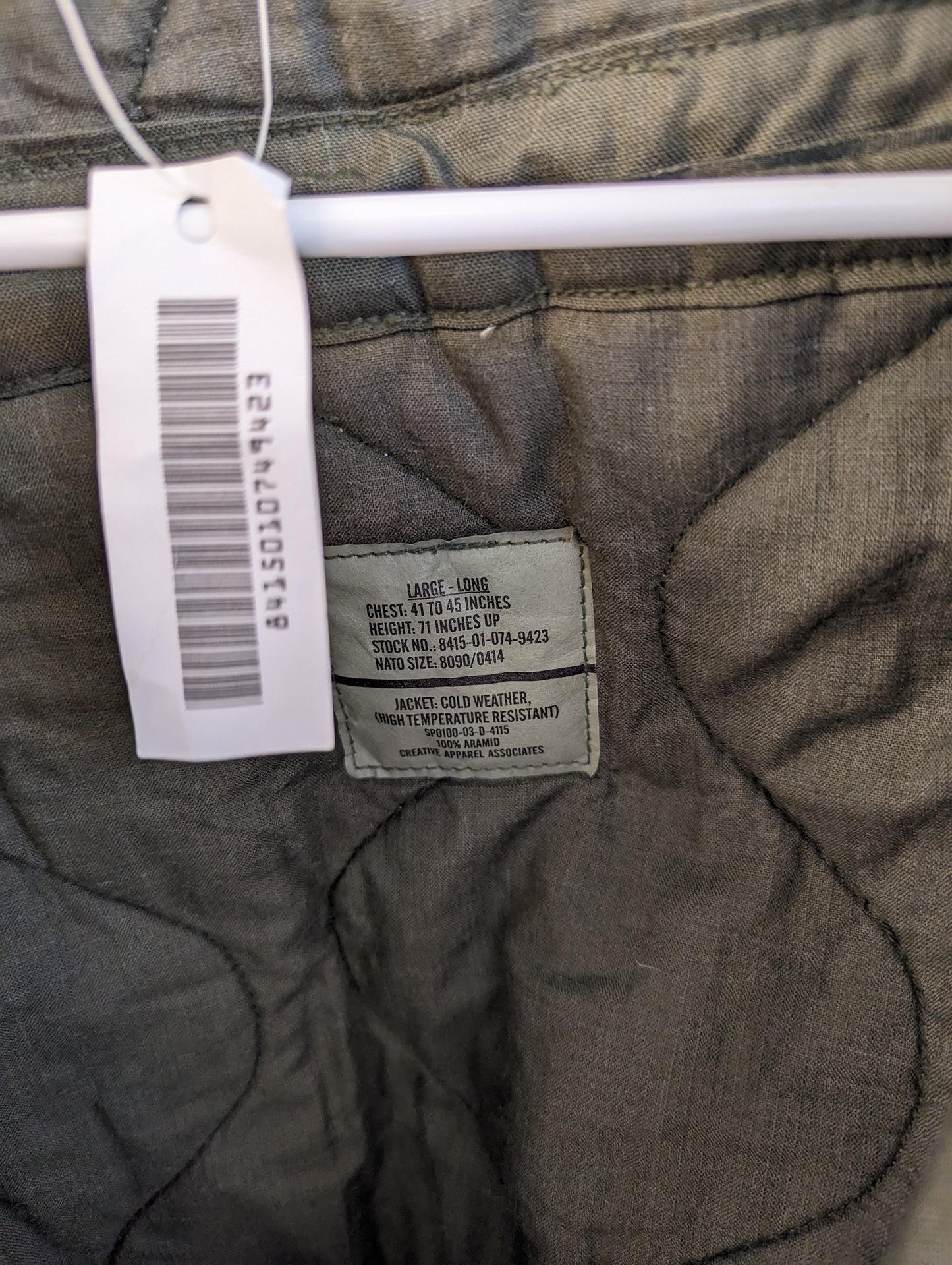 A close-up photograph of the U.S. Military issue tag sewn into the quilted CVC jacket, clearly displaying the size 'LARGE - LONG', the National Stock Number (NSN), and care instructions.