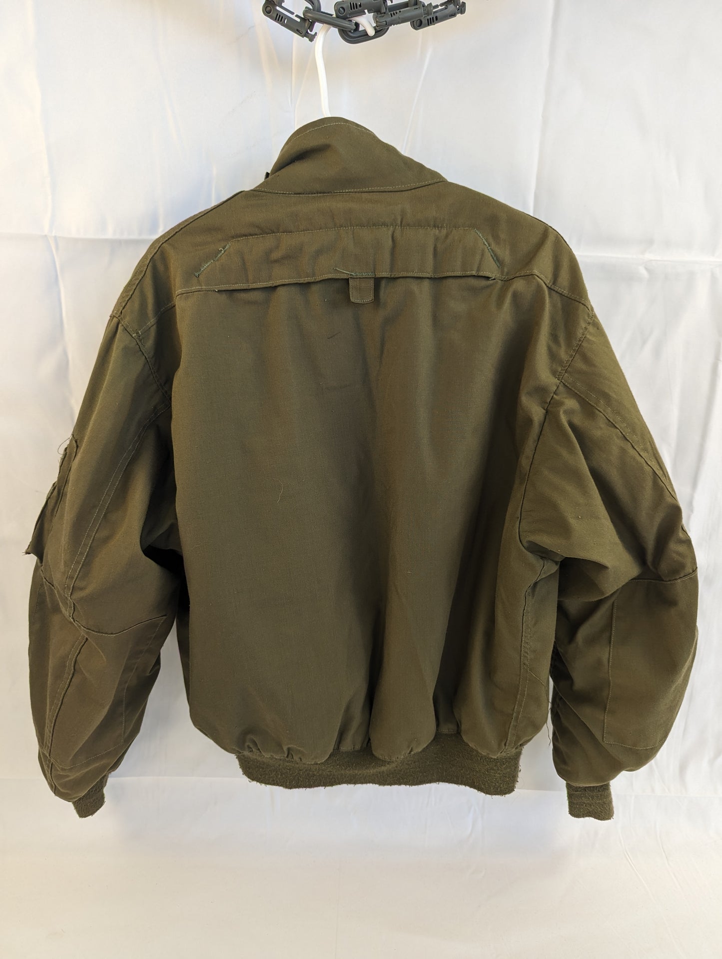 A back view of a U.S. Military Cold Weather Combat Vehicle Crew (CVC) jacket in olive green, featuring a ribbed waistband and a horizontal drag handle across the upper back, hanging on a white background.