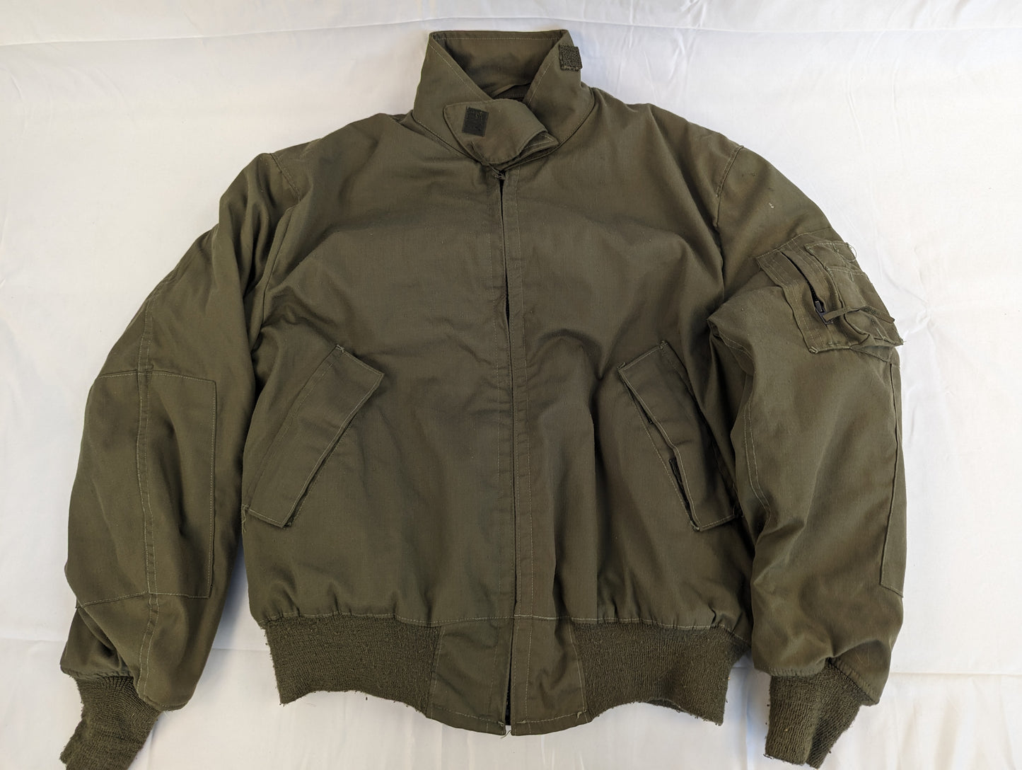 A full front view of a U.S. Military Cold Weather Combat Vehicle Crew (CVC) jacket in olive green, featuring a zippered front, ribbed cuffs, and angled chest pockets, laid flat on a white background.