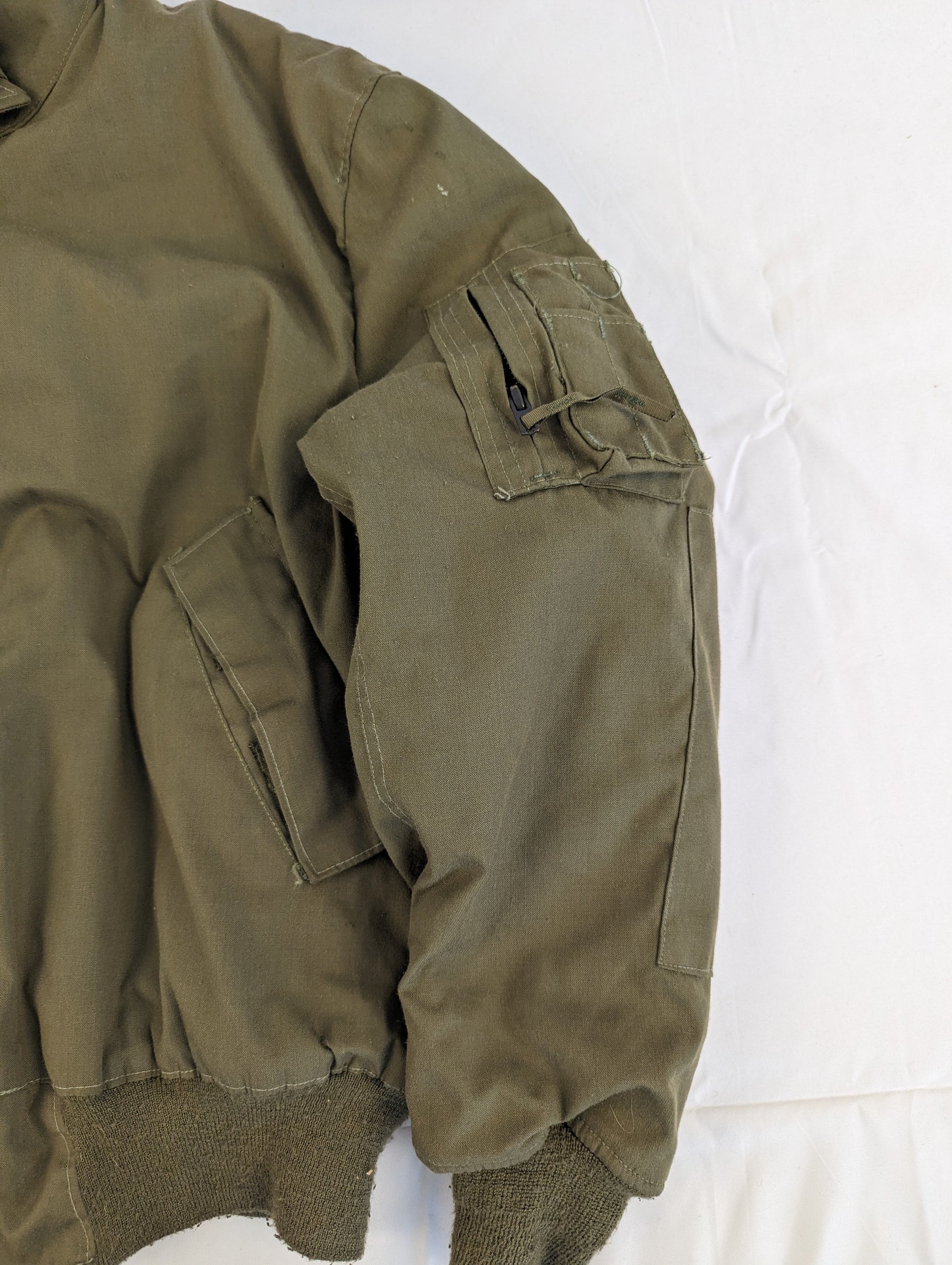 A close-up view of the left shoulder and sleeve pocket of a U.S. Military Cold Weather CVC jacket in olive green, highlighting the zippered utility pocket and ribbed cuff.