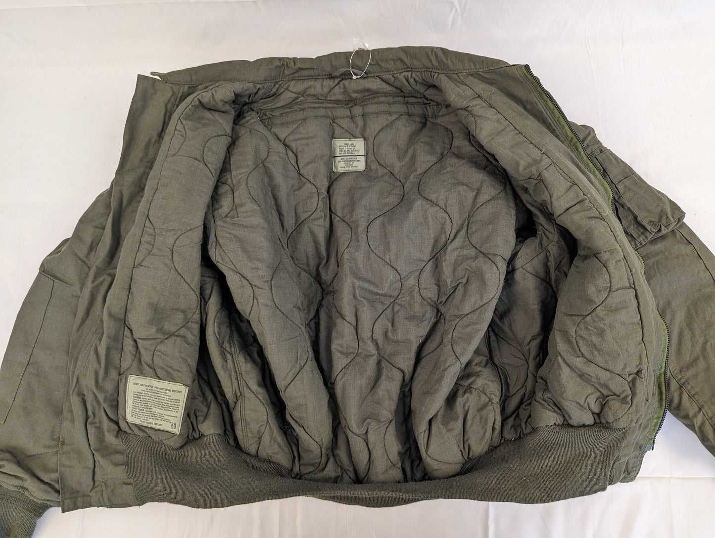 A back view of the U.S. Military CVC jacket turned inside-out, displaying the quilted olive green liner, internal tags, and the ribbed collar and waistband.