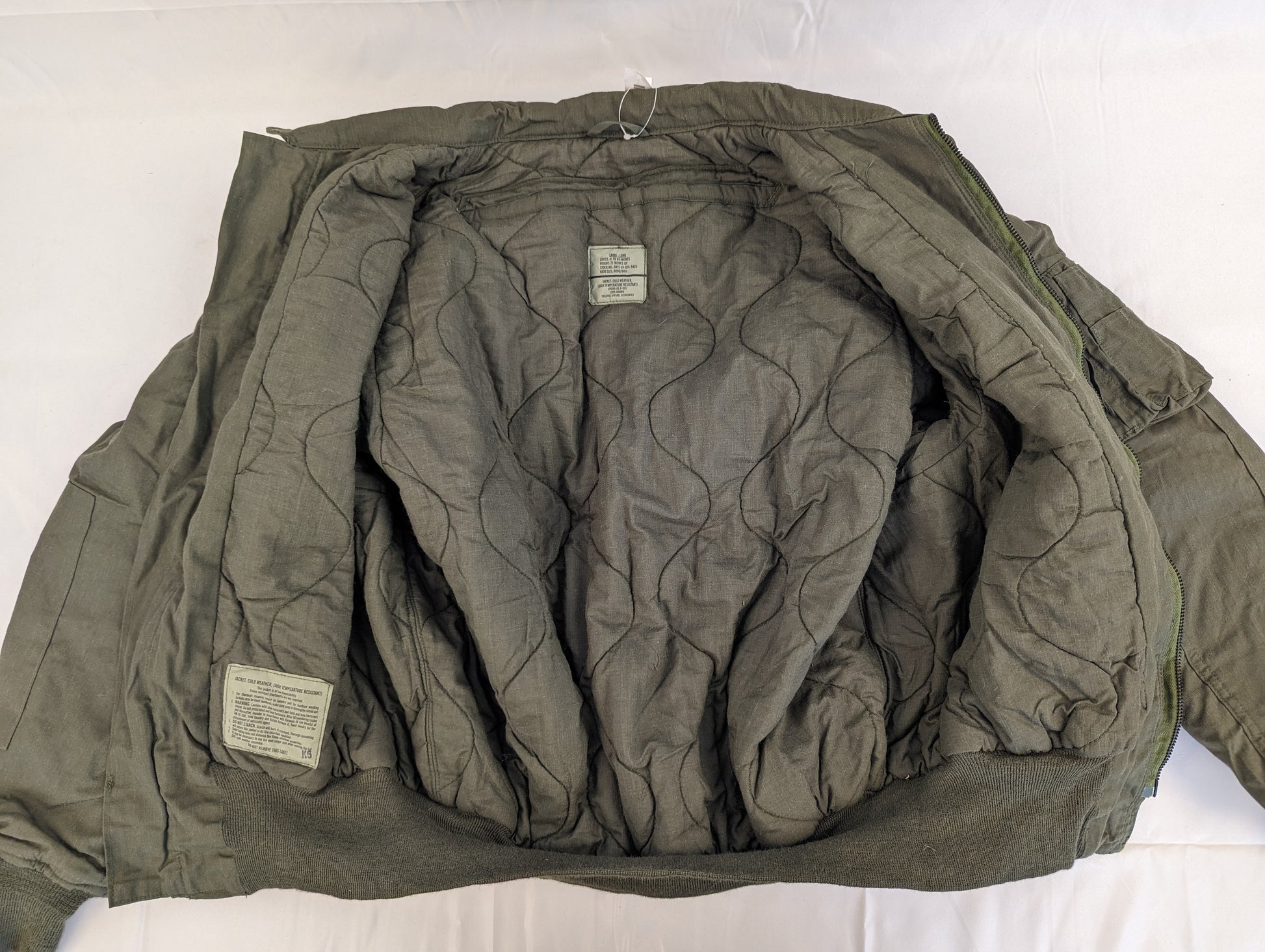A back view of the U.S. Military CVC jacket turned inside-out, displaying the quilted olive green liner, internal tags, and the ribbed collar and waistband.