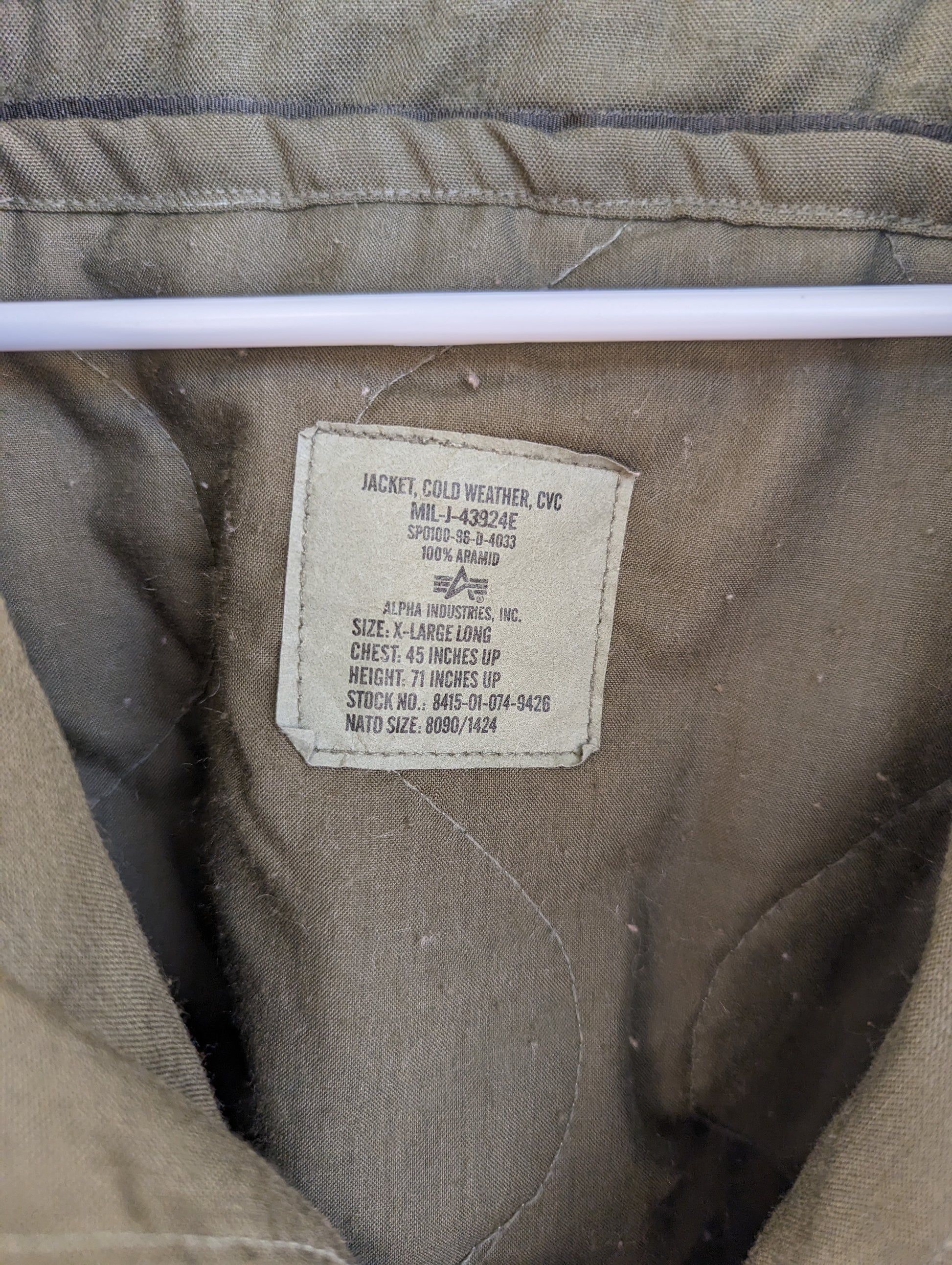 A close-up photograph of the U.S. Military issue tag sewn into the quilted CVC jacket, clearly displaying the size 'X-LARGE LONG', the National Stock Number (NSN), and the contract number.
