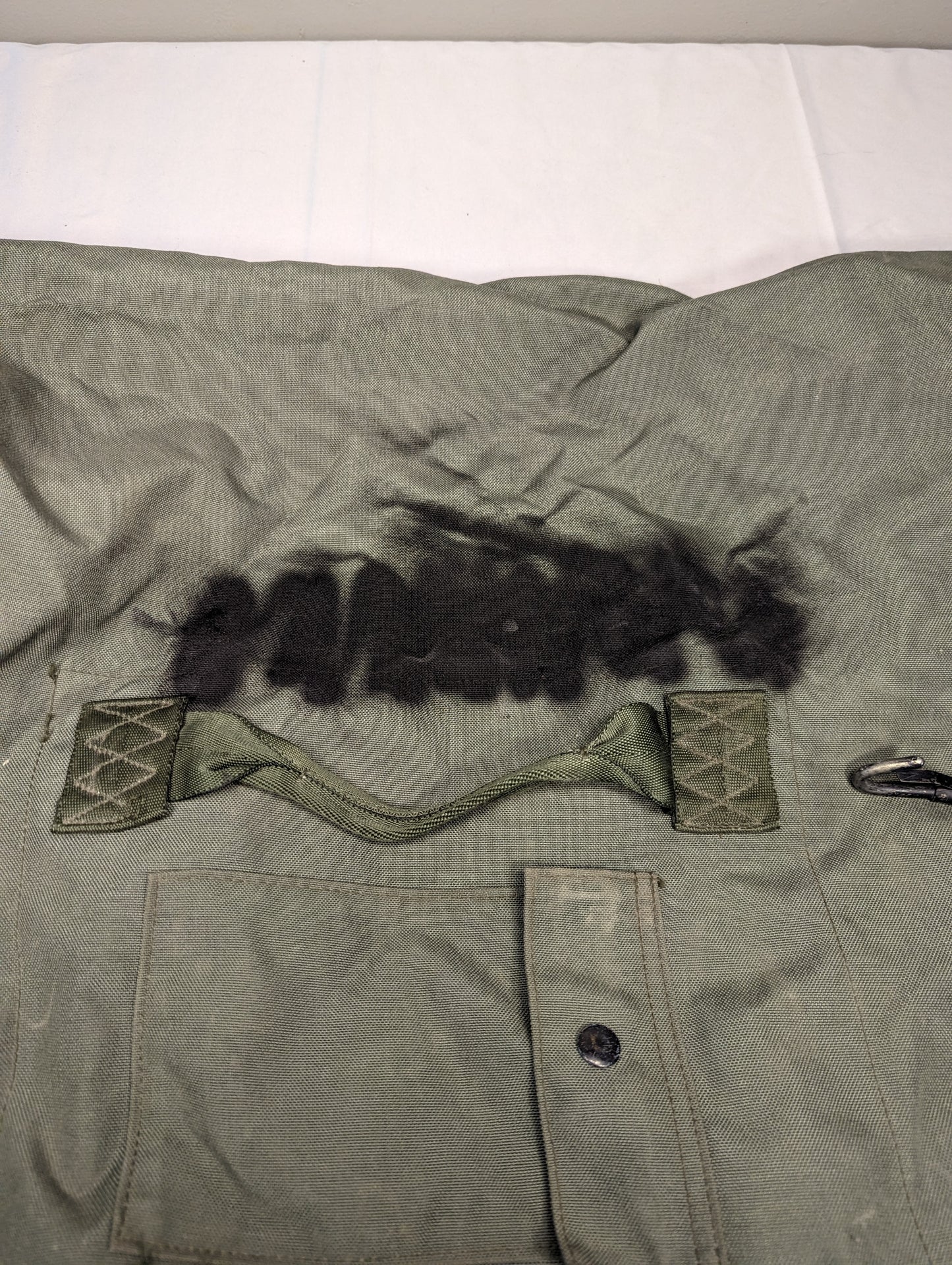 A close-up view of the wrinkled olive green fabric on a U.S. Military Duffel Bag, featuring a webbing carry handle stitched over a patch of thick black synthetic fur.