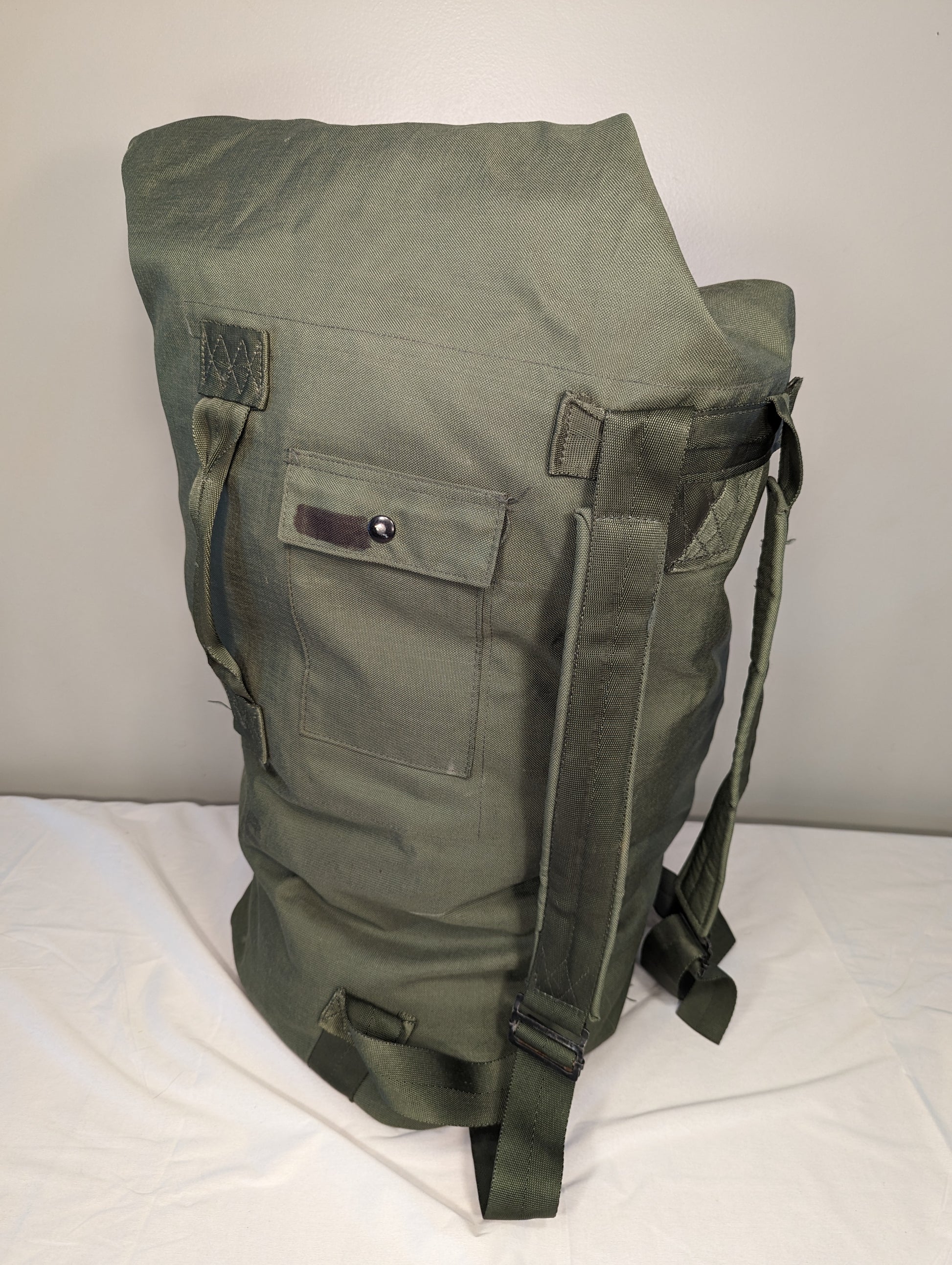 A large, olive green U.S. Military Surplus Duffel Bag, heavily compressed and standing upright, featuring thick webbing straps, side zipper closure, and a rugged, wrinkled nylon texture.