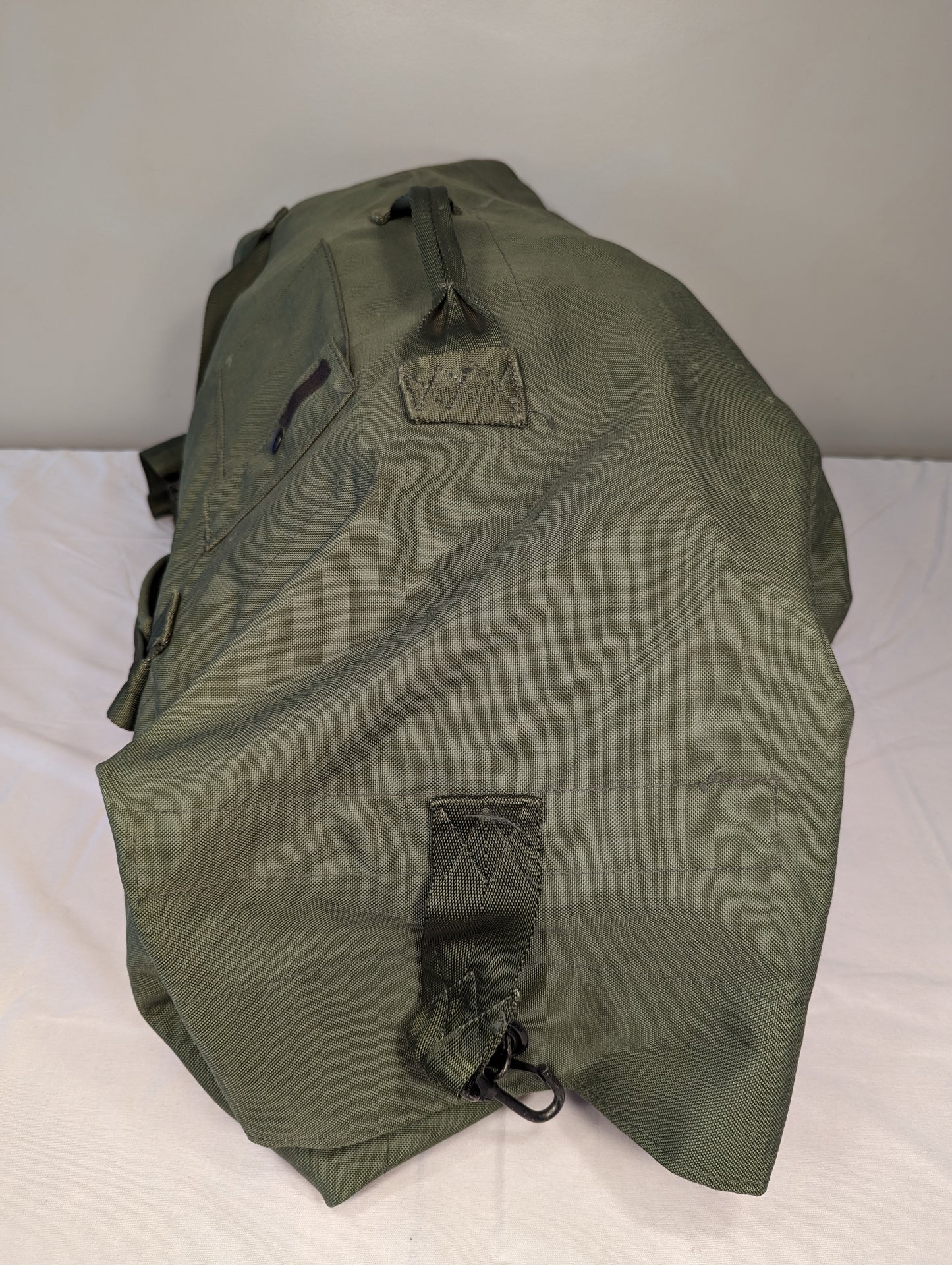 A large, olive green U.S. Military Surplus Duffel Bag laid horizontally, highlighting the heavy-duty nylon texture, webbing compression straps, and carrying handles.