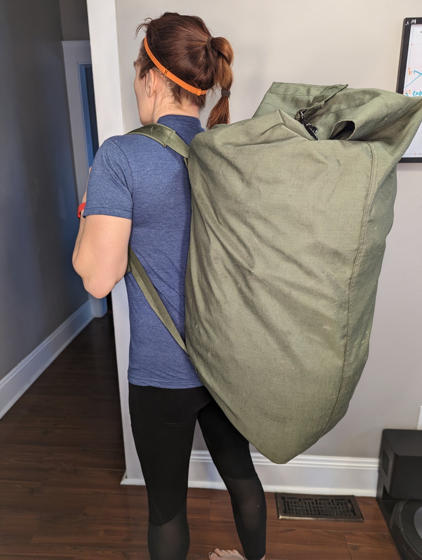 A woman standing with her back to the camera, carrying a very large, olive green U.S. Military Surplus Duffel Bag slung over her shoulder, demonstrating the bag's immense size and carry method.