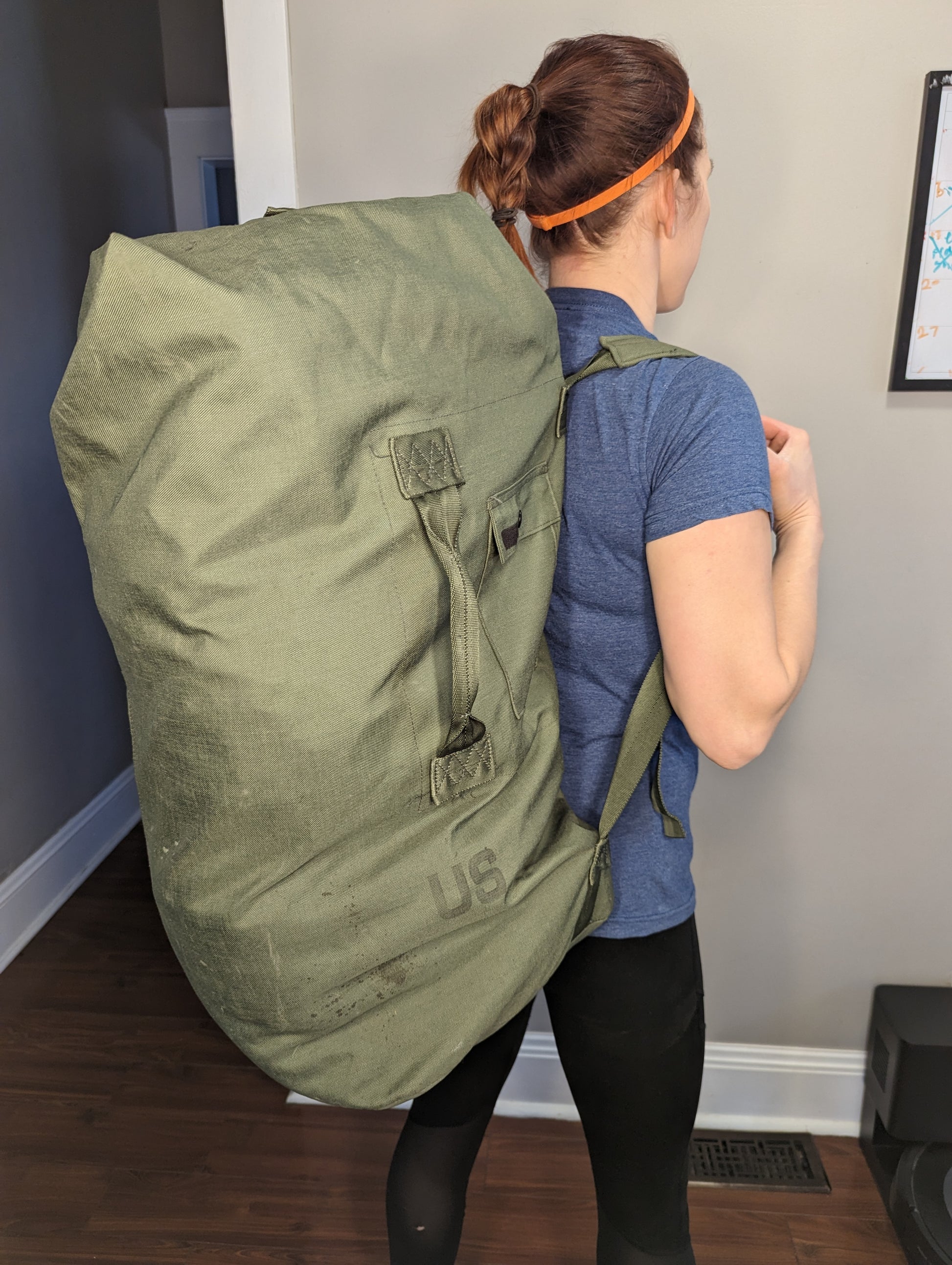 A woman viewed from the back carrying a massive olive green U.S. Military Duffel Bag slung over her shoulders, demonstrating the large size and high capacity of the cylindrical bag.
