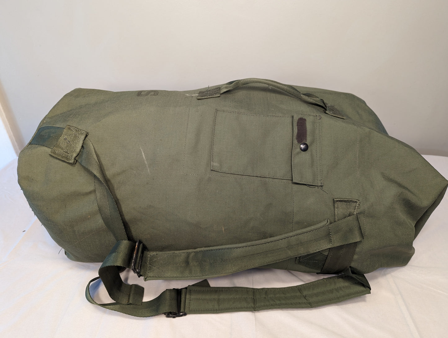 A large U.S. Military Surplus Duffel Bag in olive green, lying flat, featuring heavy-duty canvas, thick webbing straps, a side closure snap, and a prominent stenciled 'US' mark.