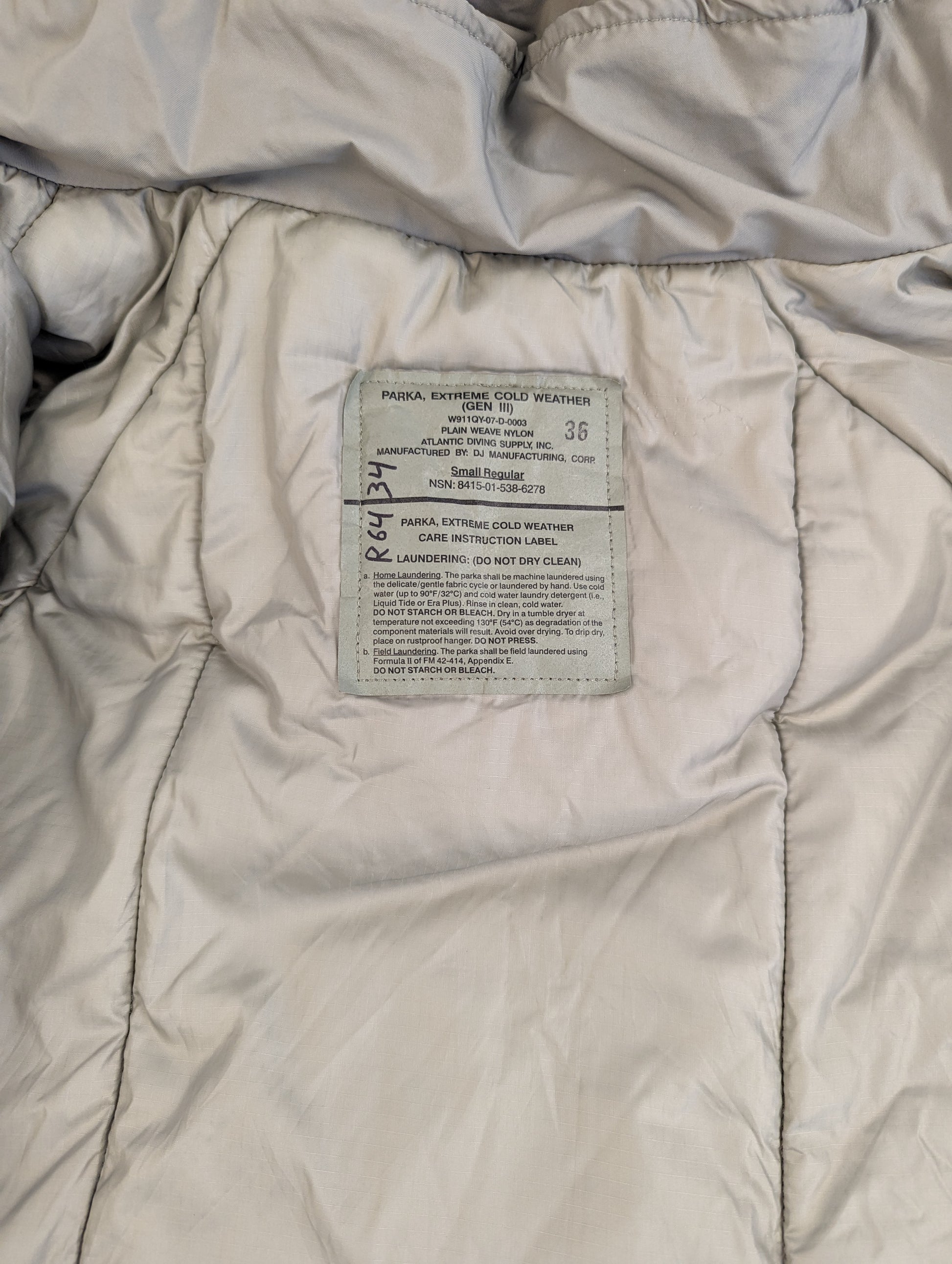 A close-up photograph of the large white U.S. Military issue tag sewn into the quilted Extreme Cold Weather Parka, displaying the product name, DLA information, and detailed care instructions.