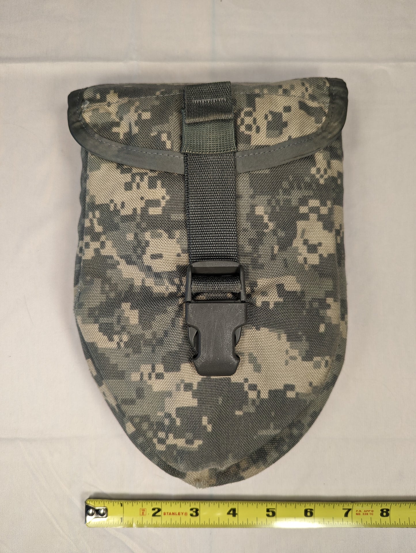 A spade-shaped U.S. Military Surplus Entrenching Tool Carrier pouch in ACU digital camouflage, laid flat with a black quick-release buckle, and a measuring tape across the base showing its approximate 9-inch length.