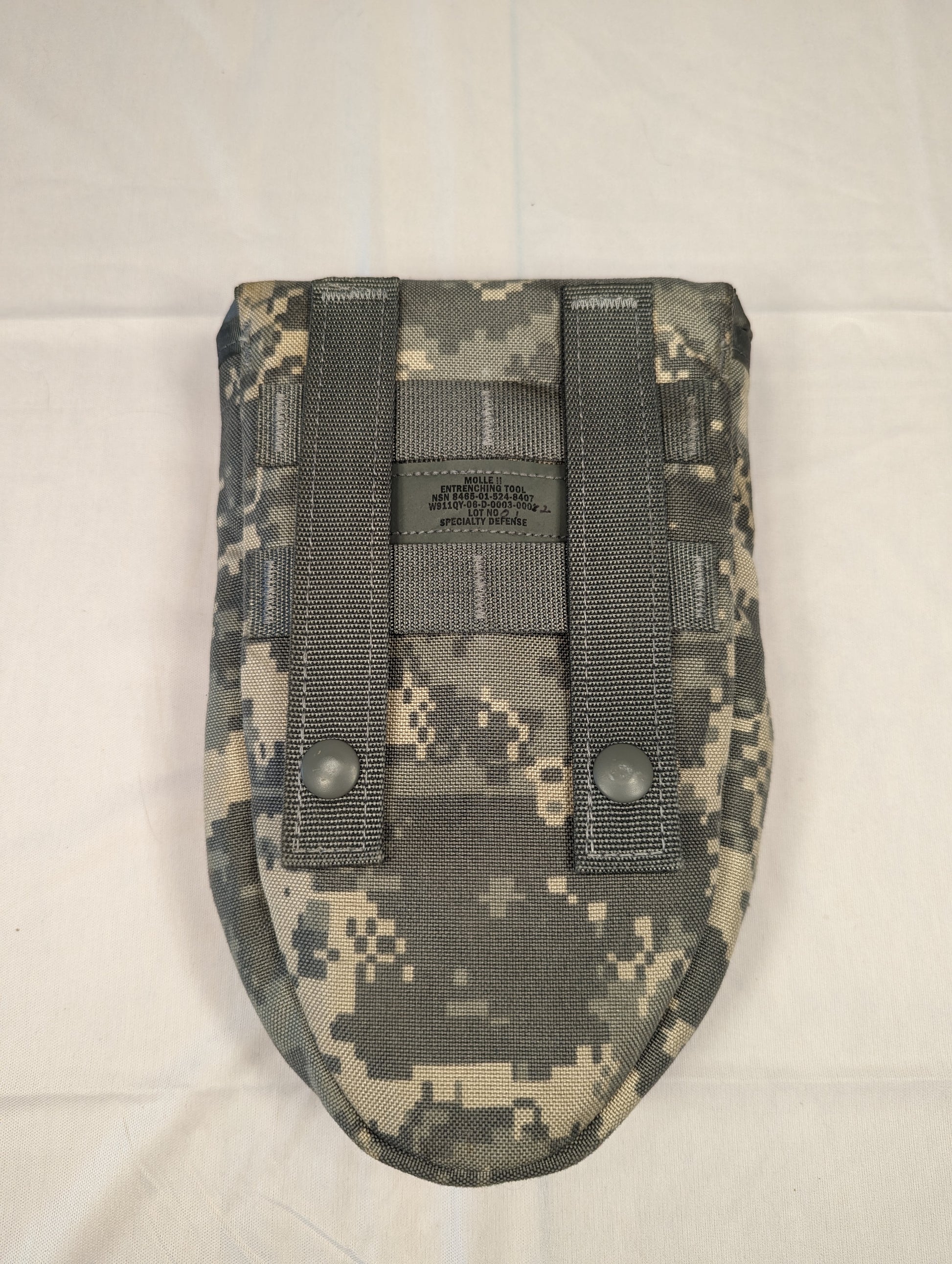 A back view of a U.S. Military Surplus Entrenching Tool Carrier pouch in ACU digital camouflage, clearly displaying the two vertical MOLLE webbing attachment straps, metal snap buttons, and stamped issue details.