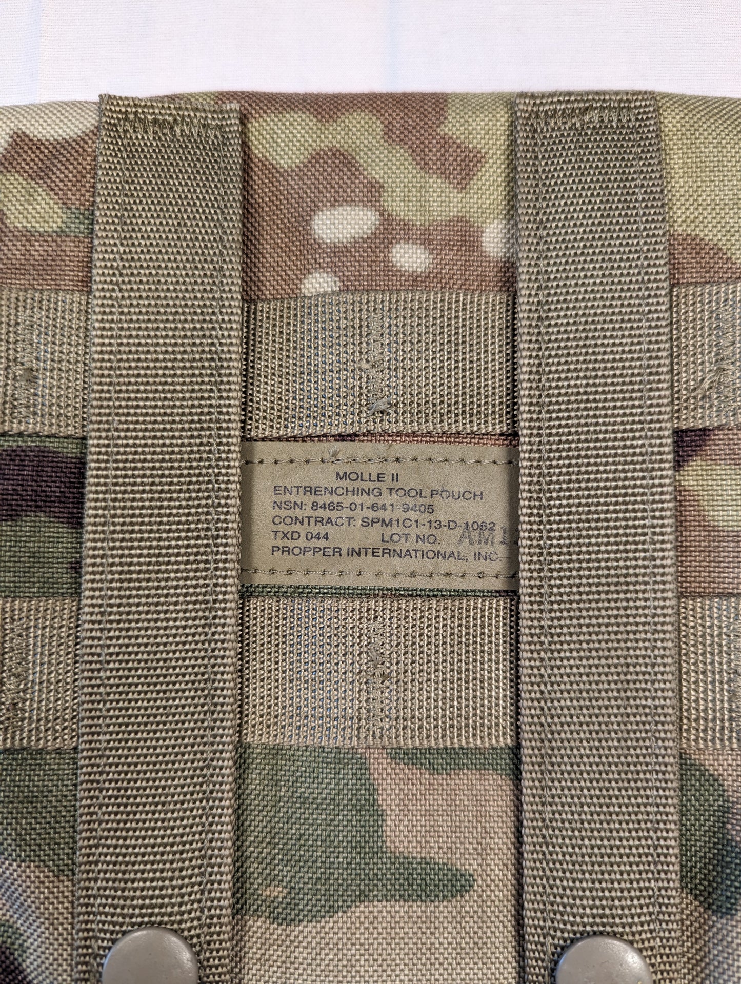 A close-up photograph of the original U.S. Military issue tag sewn into the OCP/Multicam Entrenching Tool Carrier pouch, clearly displaying the product name, NSN, and manufacturer.