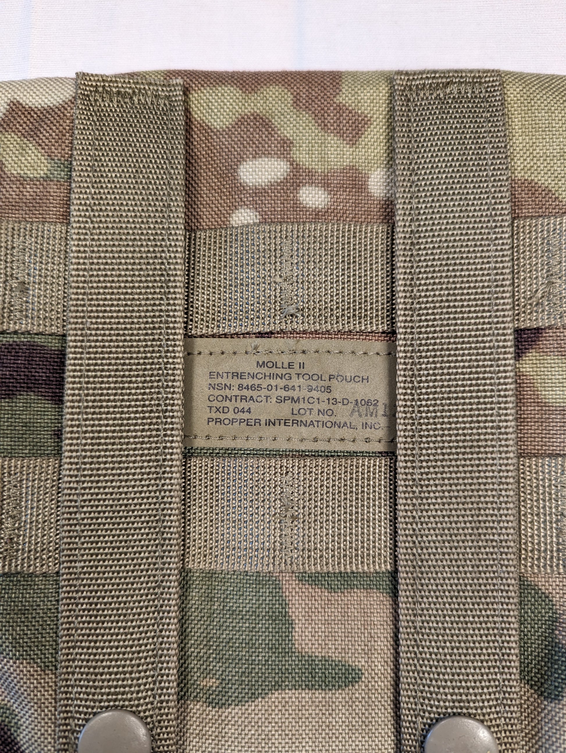 A close-up photograph of the original U.S. Military issue tag sewn into the OCP/Multicam Entrenching Tool Carrier pouch, clearly displaying the product name, NSN, and manufacturer.