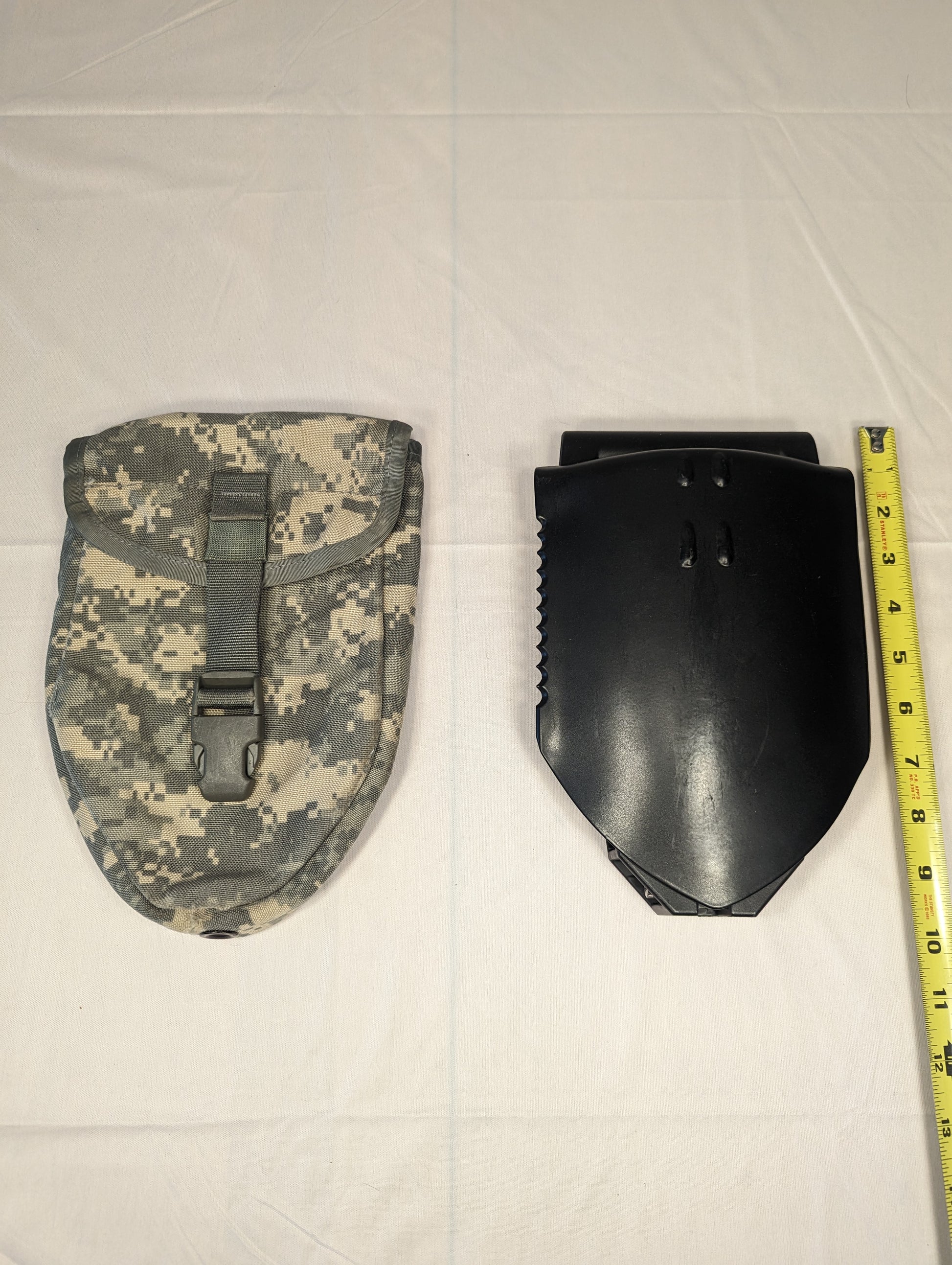 A folded black U.S. Military Entrenching Tool (E-Tool) with a serrated steel blade, displayed next to its ACU digital camouflage carrying pouch and a yellow tape measure demonstrating its compact length.