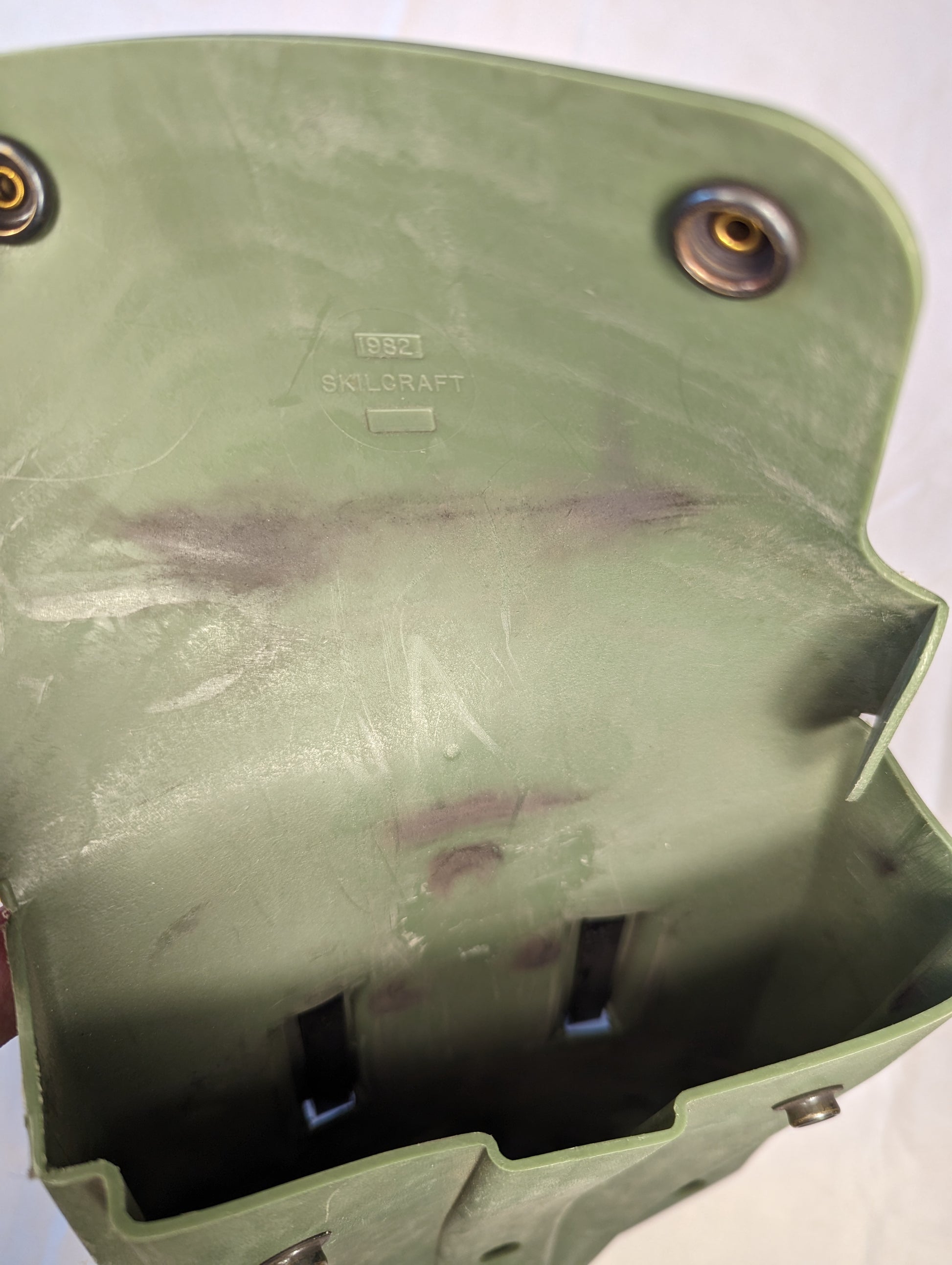 A close-up view of the open interior of a U.S. Military Surplus plastic entrenching tool carrier case in olive drab green, displaying the embossed '1982' and 'SKILCRAFT' manufacturer markings and visible signs of field use.