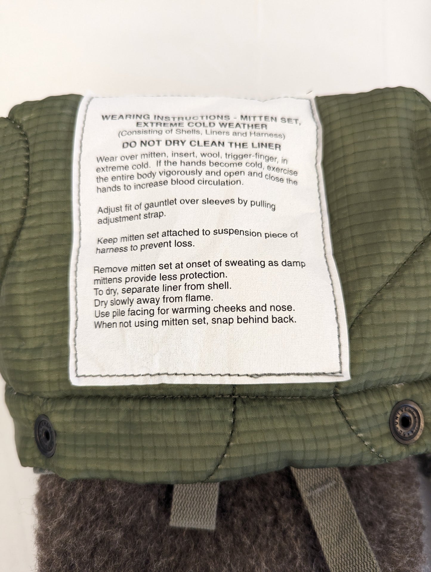 A close-up photograph of the white instruction label sewn into the U.S. Military Extreme Cold Weather Mitten set, displaying detailed wearing and removal instructions for extreme cold, including advice on sweating and drying.