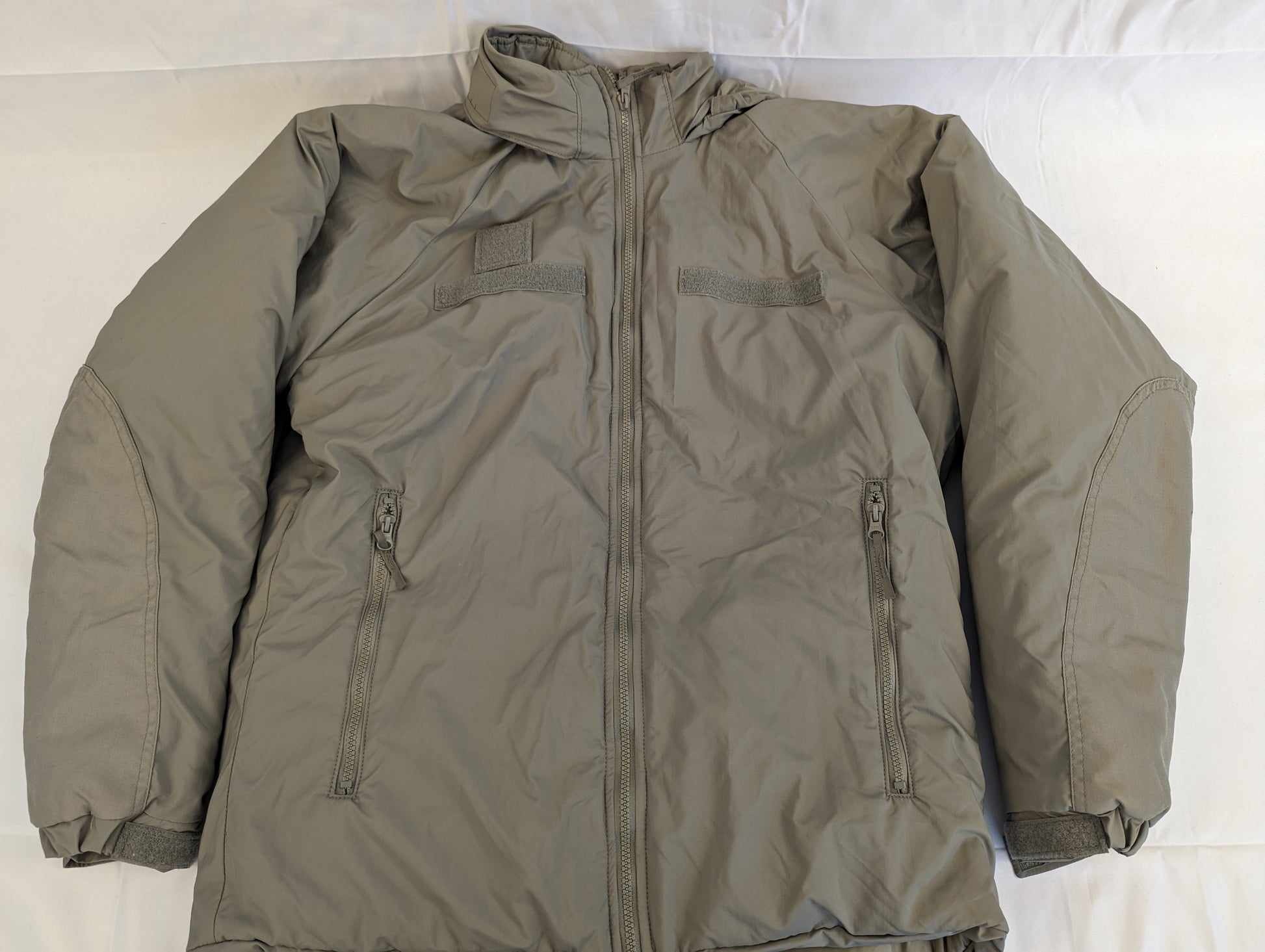 A full front view of a gray U.S. Military Extreme Cold Weather Parka laid flat, featuring a full-length zipper, a high collar, two large zippered pockets, and hook-and-loop chest patches.