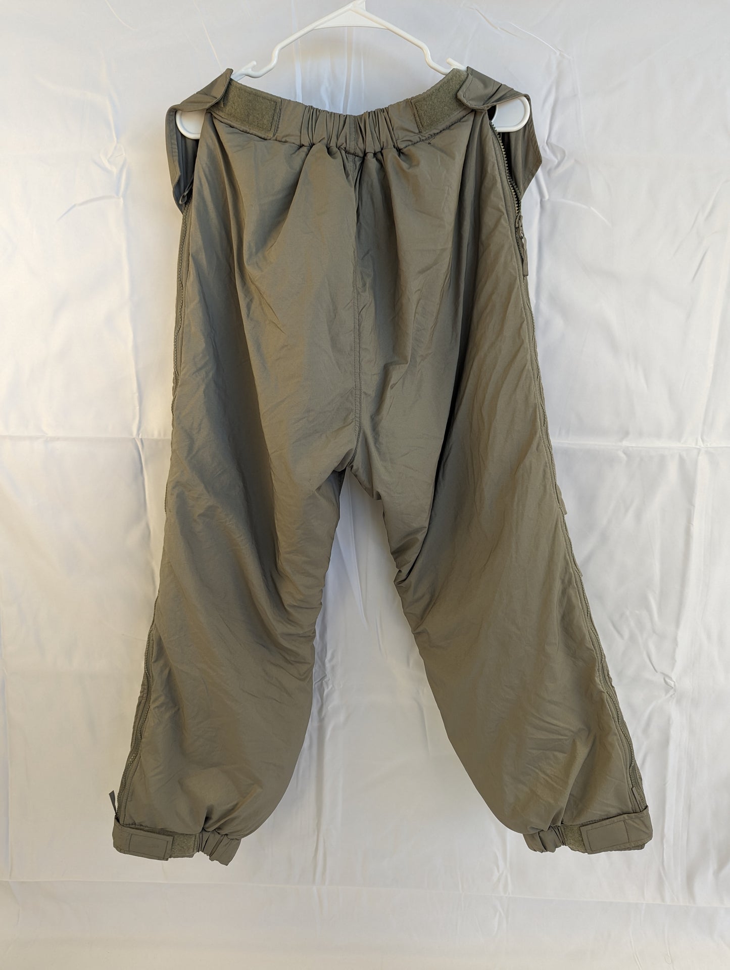A full-length back view of U.S. Military Surplus Extreme Cold Weather Trousers in gray, hanging on a white hanger, highlighting the bulky, quilted insulation and cinched ankle cuffs.