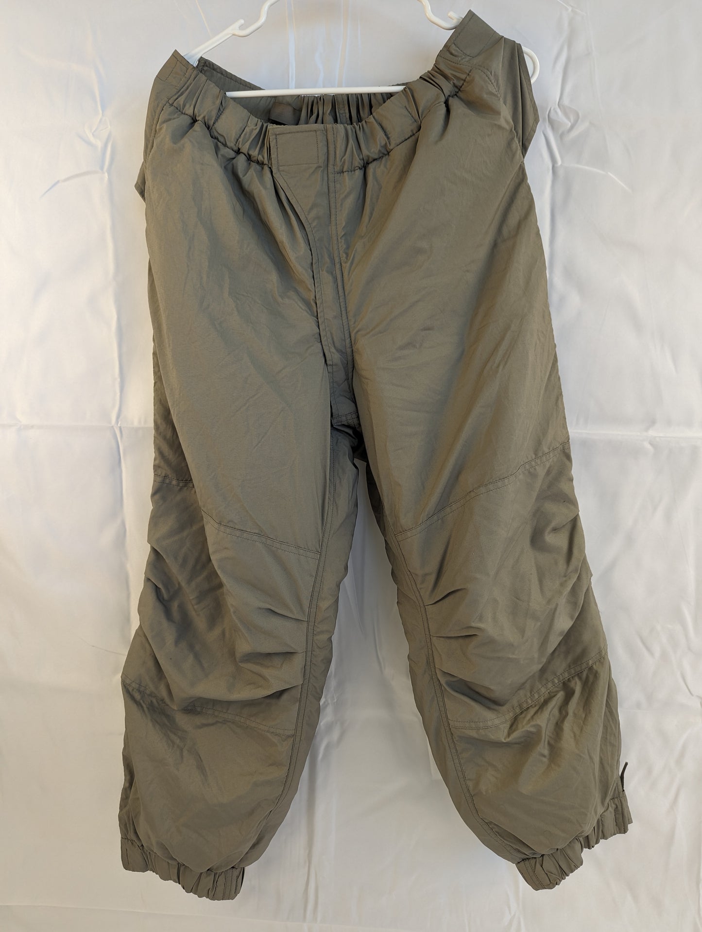 A full-length front view of a pair of U.S. Military Surplus Extreme Cold Weather Trousers in gray, hanging on a hanger, highlighting the bulky, quilted insulation, elastic waist, and cinched cuffs.