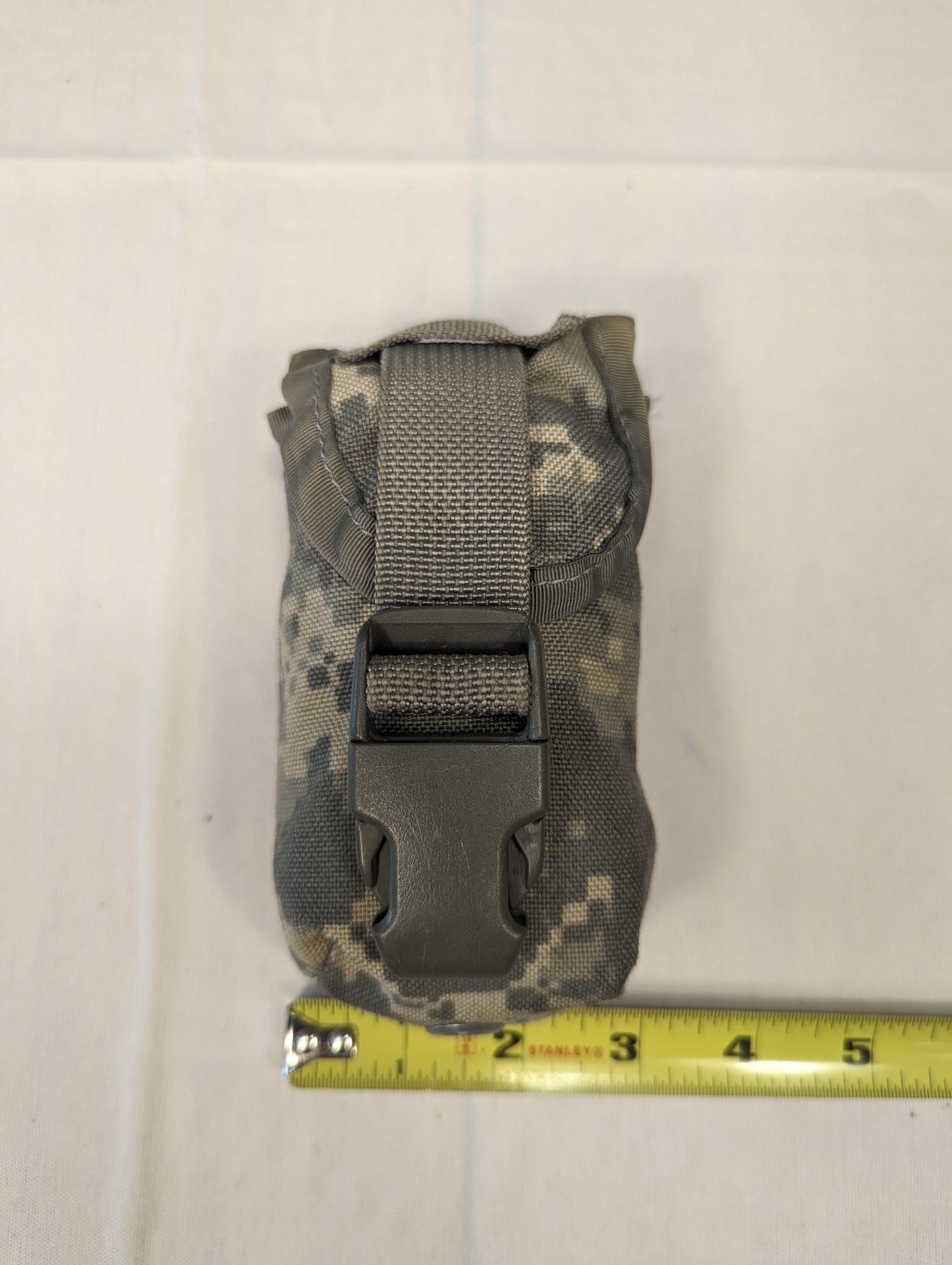 A single U.S. Military Surplus FG Pouch in ACU digital camouflage, standing upright, secured by a black quick-release buckle and webbing, with a measuring tape positioned below to demonstrate its compact size.