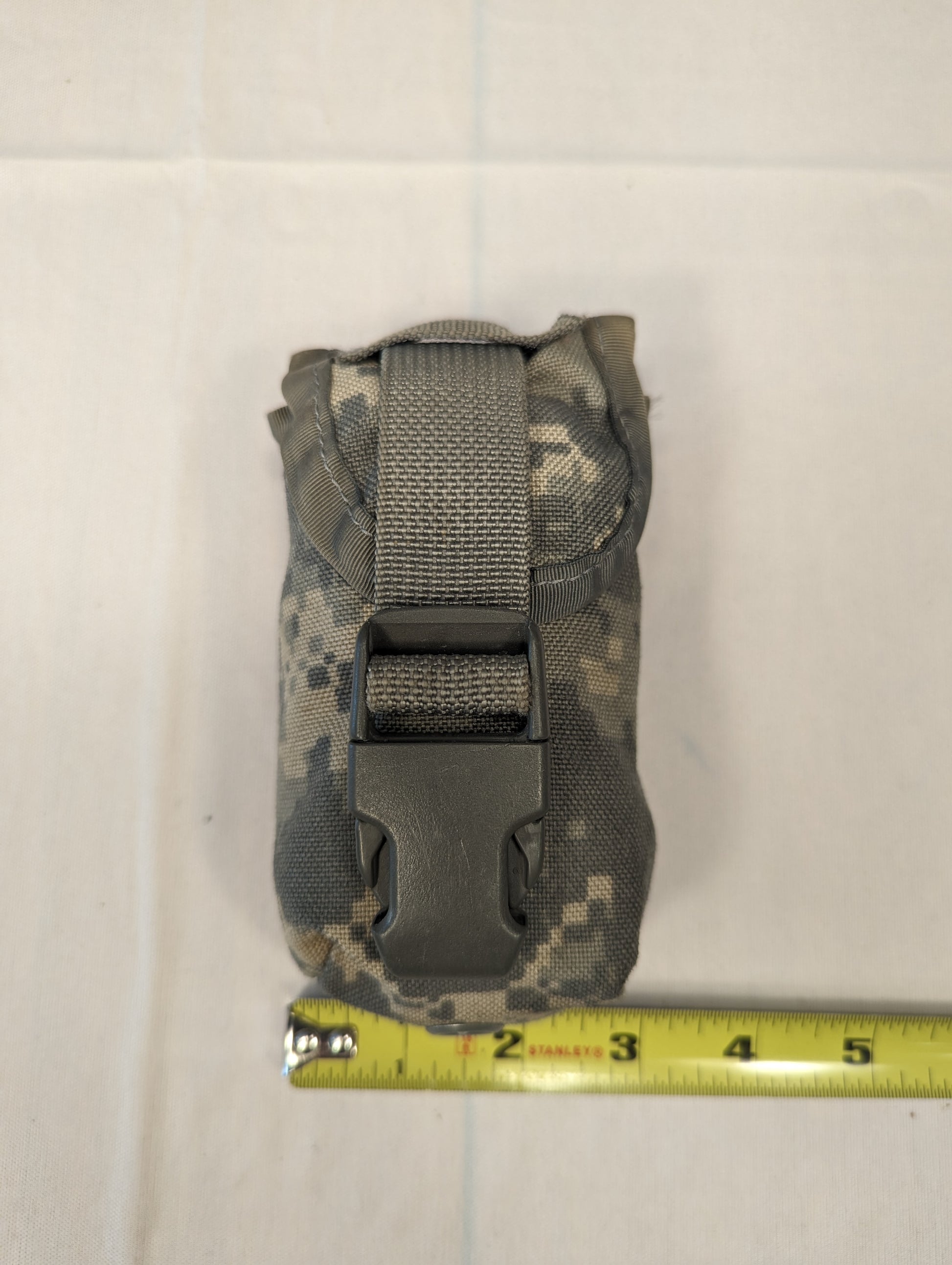 A single U.S. Military Surplus FG Pouch in ACU digital camouflage, standing upright, secured by a black quick-release buckle and webbing, with a measuring tape positioned below to demonstrate its compact size.