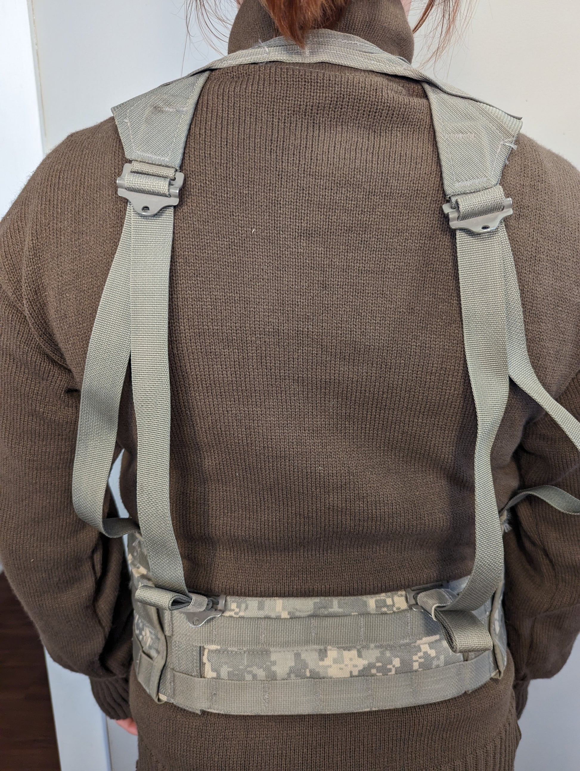 A rear view of a person wearing a U.S. Military Surplus Fighting Load Carrier (FLC) harness, highlighting the padded gray shoulder straps and the ACU digital camouflage waist belt fastened around the lower back.