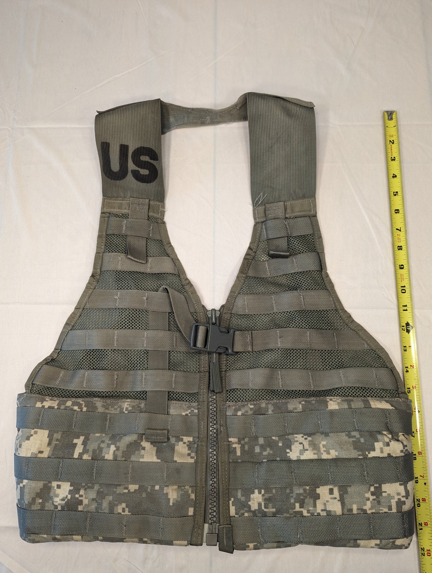 A front view of a U.S. Military Surplus Fighting Load Carrier (FLC) Vest in ACU digital camouflage, laid flat, featuring a central zipper, extensive horizontal MOLLE webbing, a stenciled 'US' mark, and a tape measure showing its vertical height.