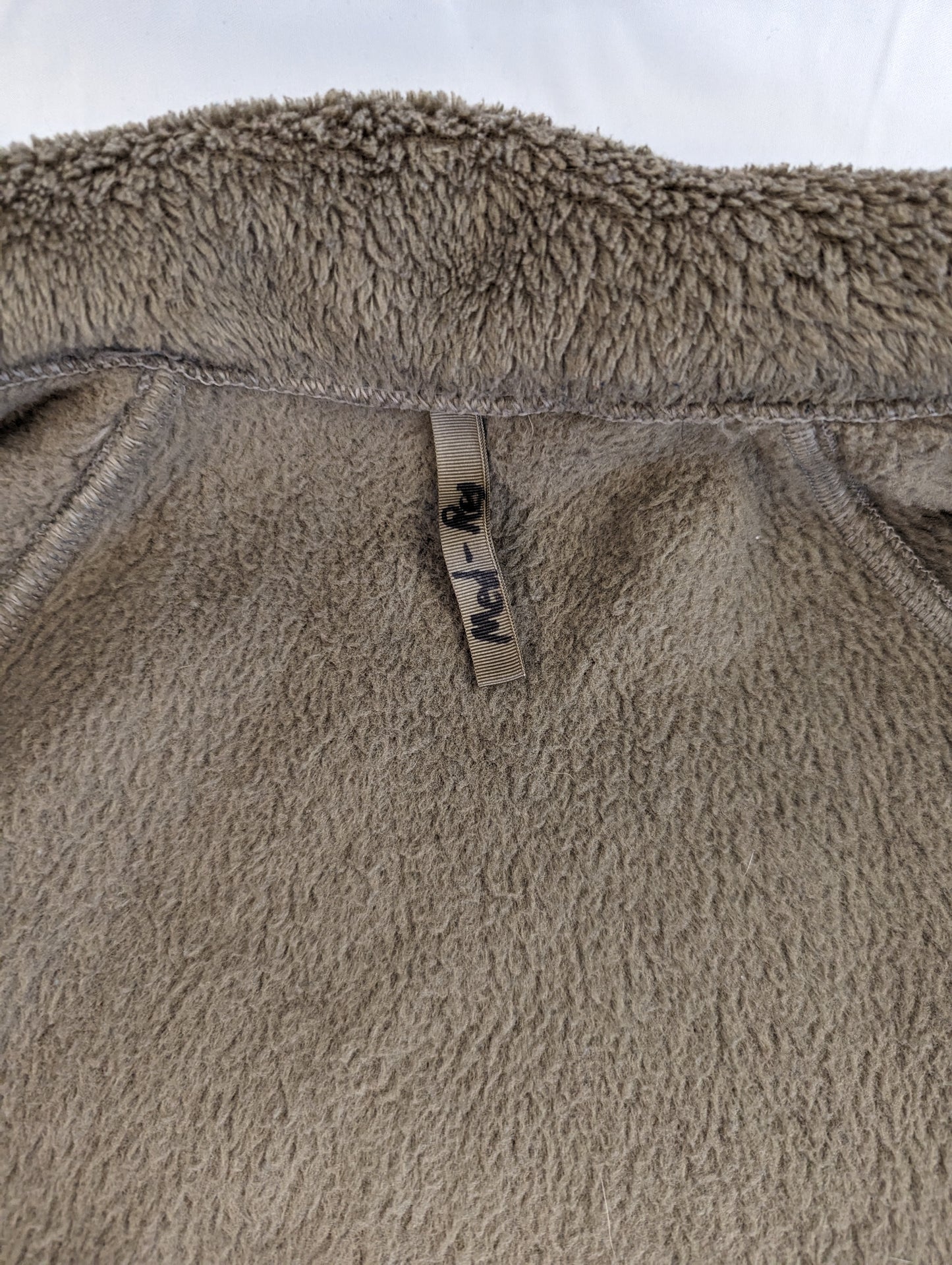 A very close-up detail shot of the high-pile Coyote Brown fleece material of the cold weather jacket, showing the fuzzy texture and the woven tag near the collar.