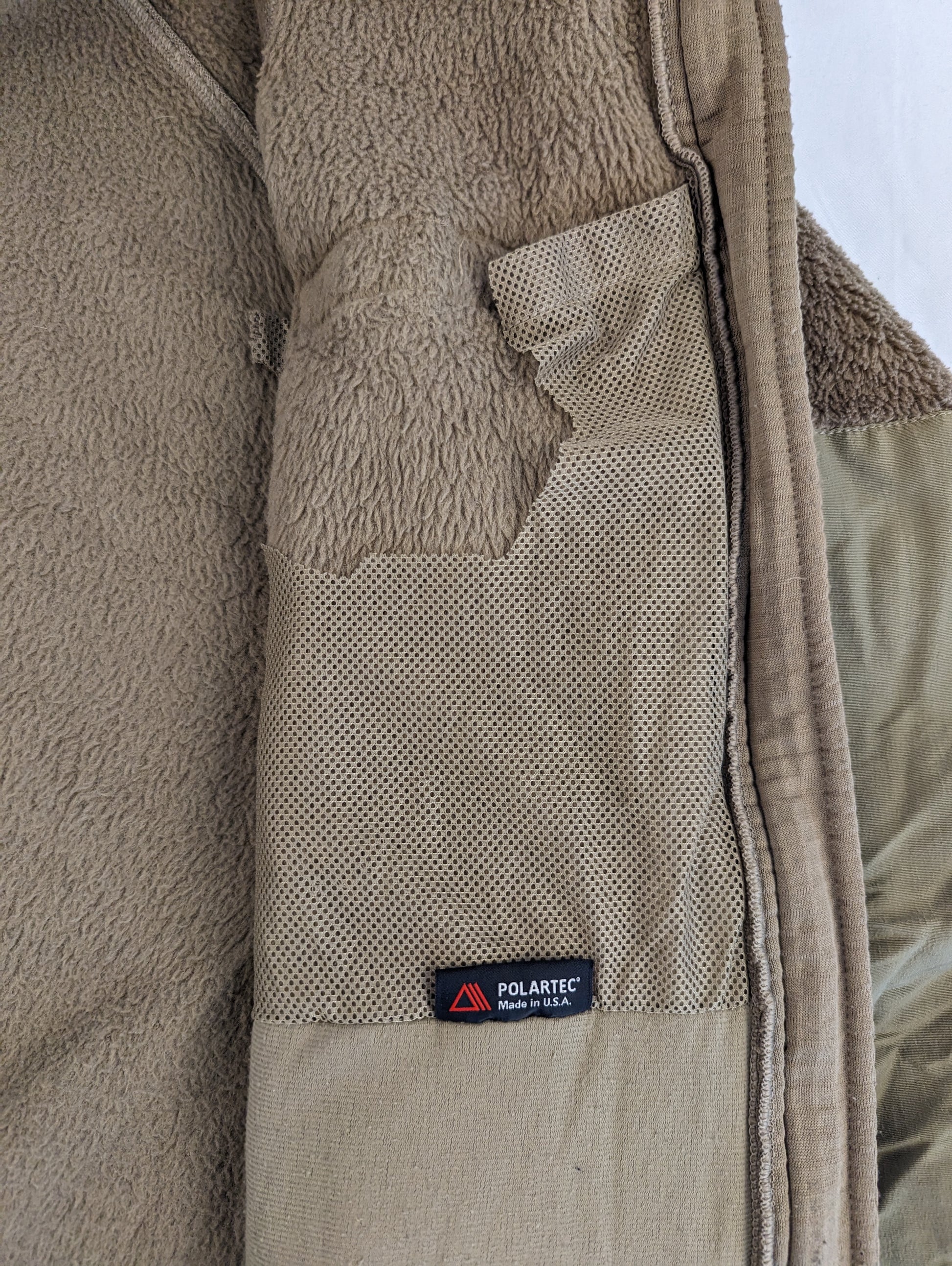 A very close-up detail shot of the interior of the Coyote Brown fleece jacket, showing the fuzzy fleece texture, a reinforced pocket flap, and the small 'POLARTEC MADE IN U.S.A.' tag.
