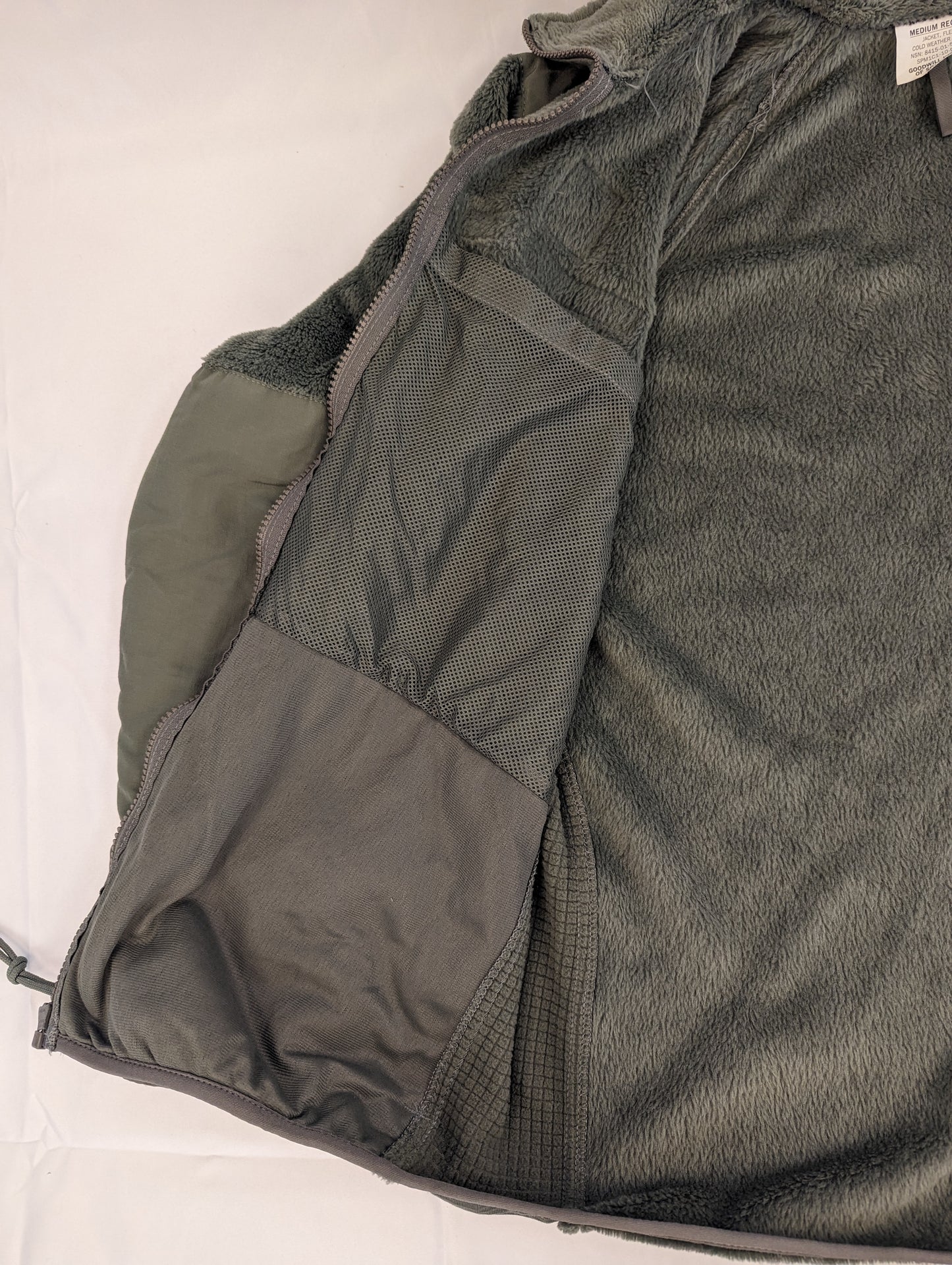 A close-up view of the interior of the U.S. Military Cold Weather Fleece Jacket in olive green, highlighting the thick fleece material, the reinforced nylon zipper placket, and the large internal mesh pocket.