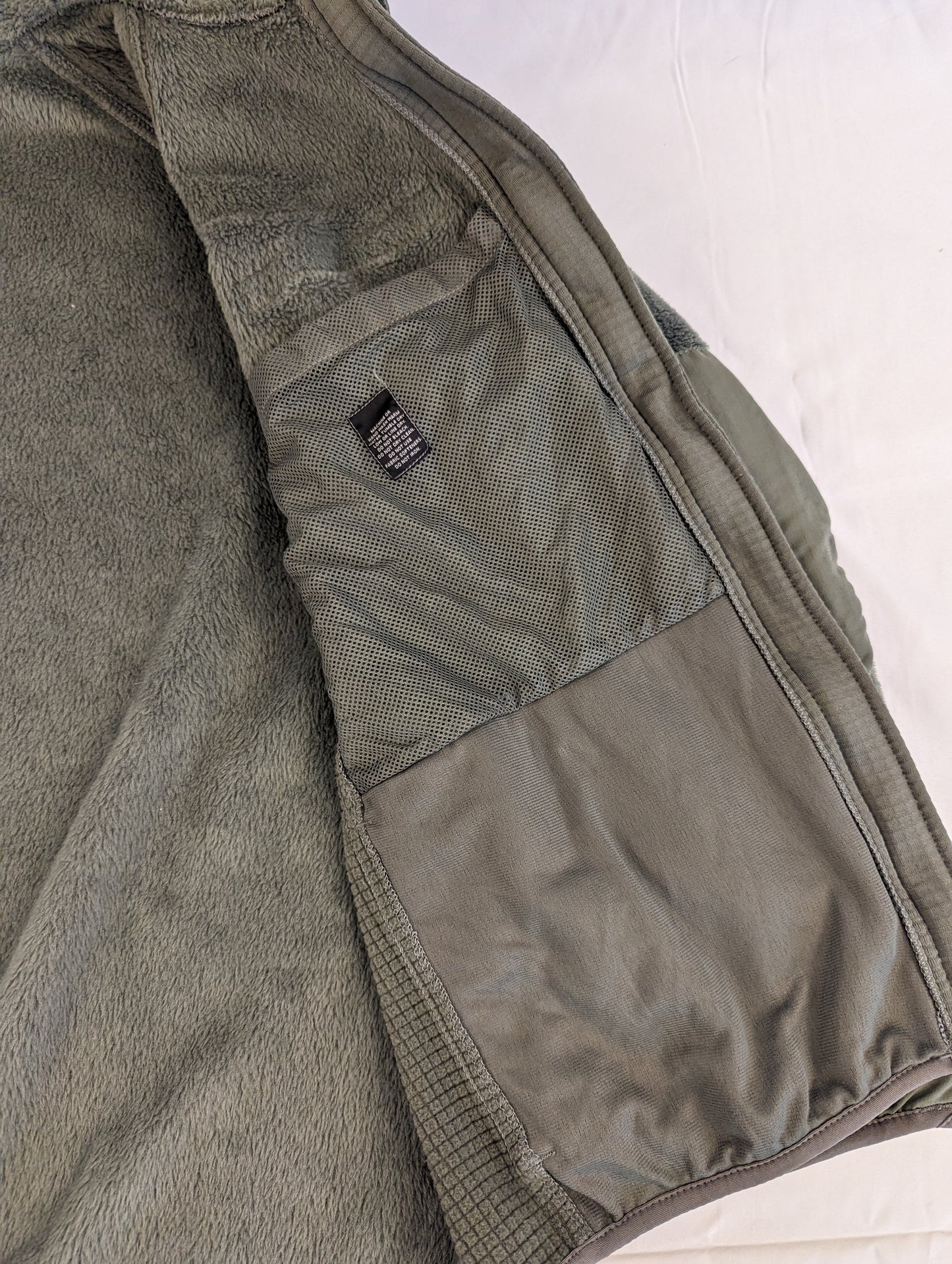 A close-up view of the interior of the U.S. Military Cold Weather Fleece Jacket in olive green, showing the fuzzy fleece texture and a large, open mesh pocket sewn into the side.