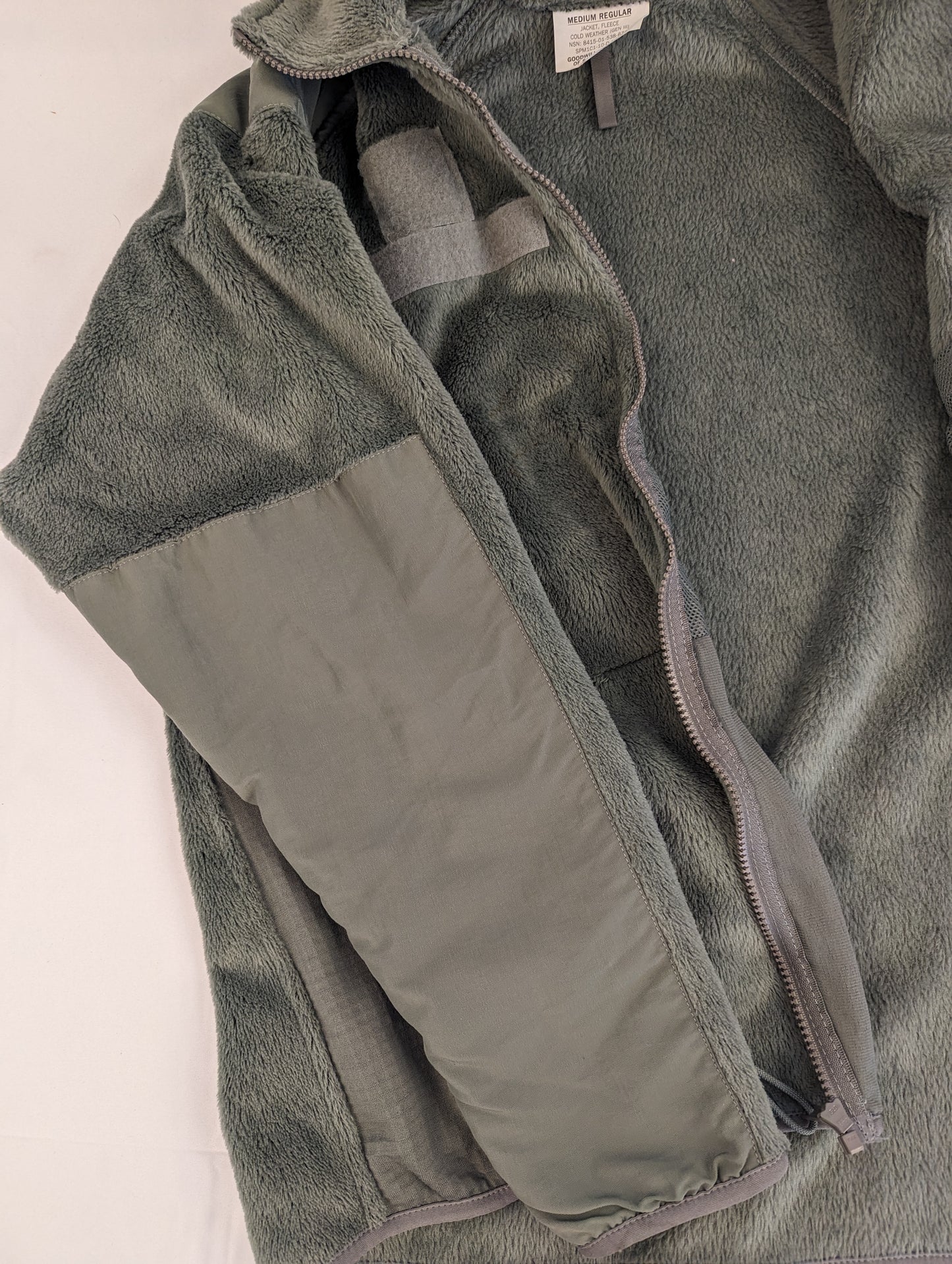 A close-up view of the left sleeve and shoulder of the olive green U.S. Military Cold Weather Fleece Jacket, highlighting the reinforced nylon elbow patch and the hook-and-loop field.
