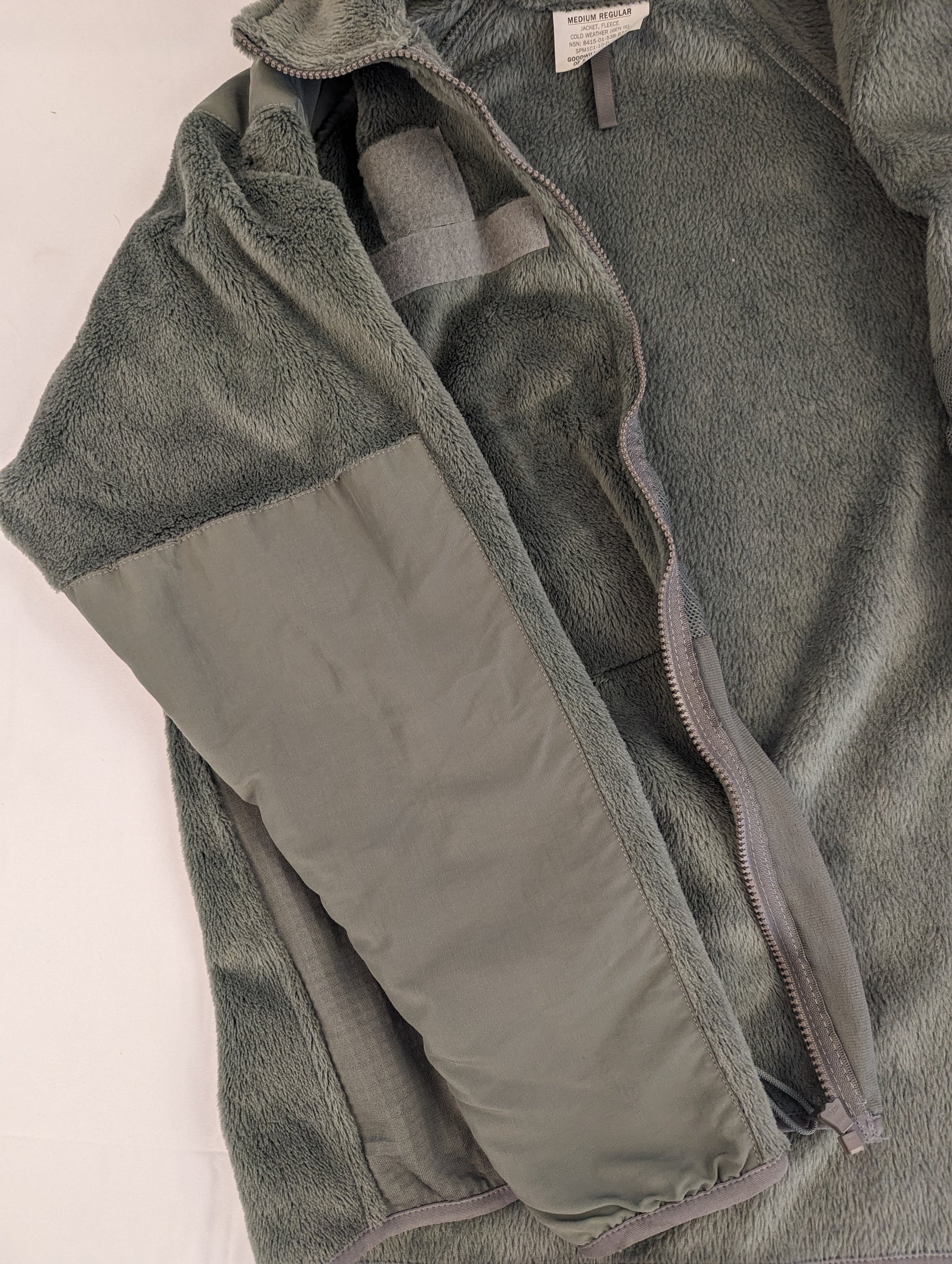 A close-up view of the left sleeve and shoulder of the olive green U.S. Military Cold Weather Fleece Jacket, highlighting the reinforced nylon elbow patch and the hook-and-loop field.