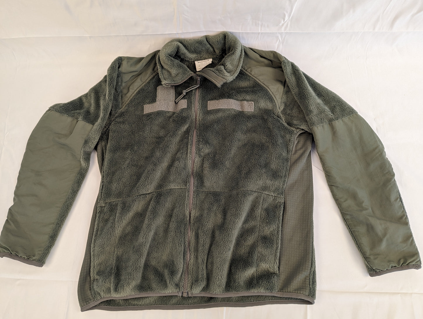 A front view of a U.S. Military Surplus Cold Weather Fleece Jacket in olive green, laid flat, featuring extensive reinforced nylon panels on the shoulders and arms, hook-and-loop patches on the chest, and breathable mesh side panels.