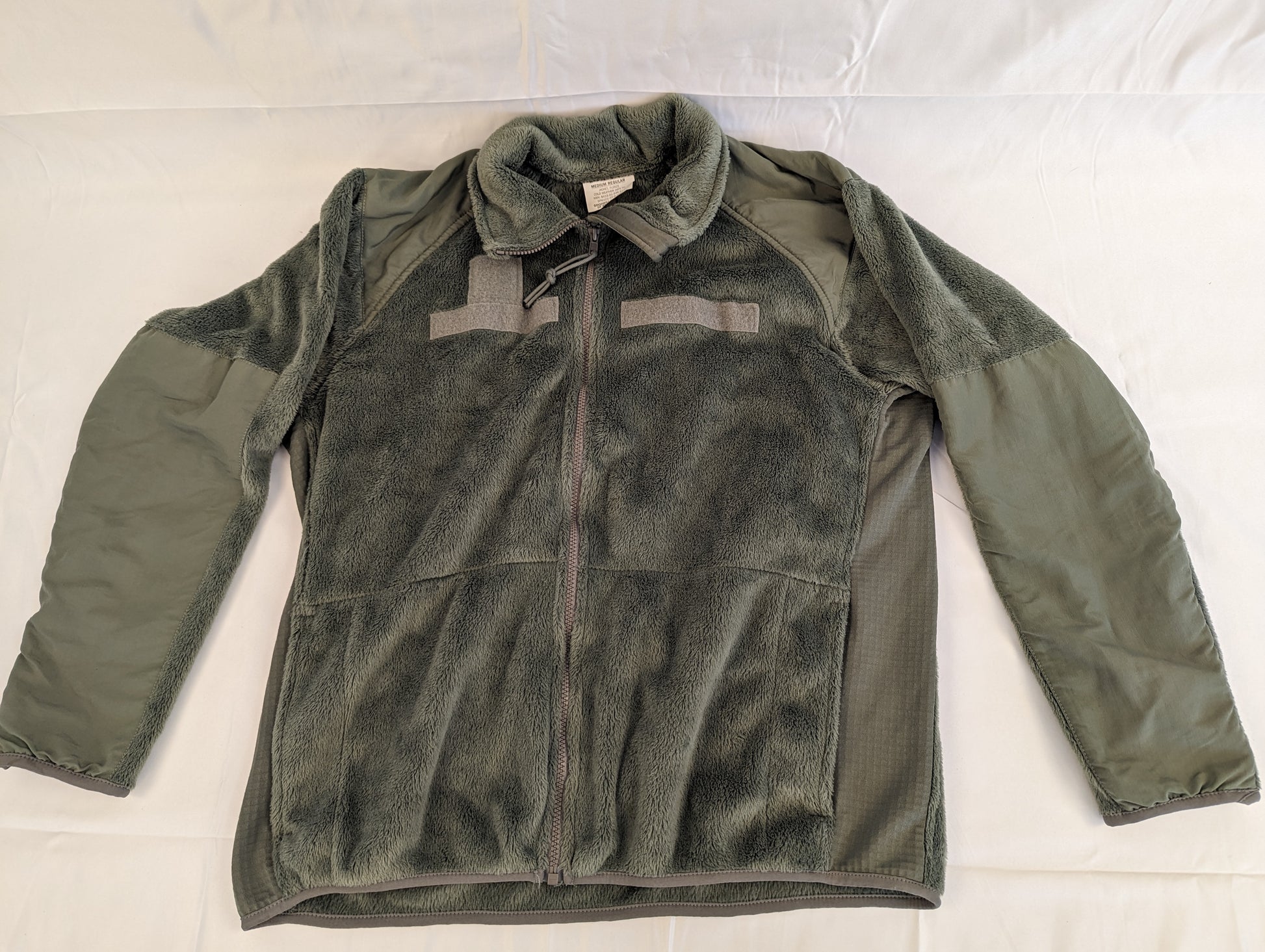 A front view of a U.S. Military Surplus Cold Weather Fleece Jacket in olive green, laid flat, featuring extensive reinforced nylon panels on the shoulders and arms, hook-and-loop patches on the chest, and breathable mesh side panels.