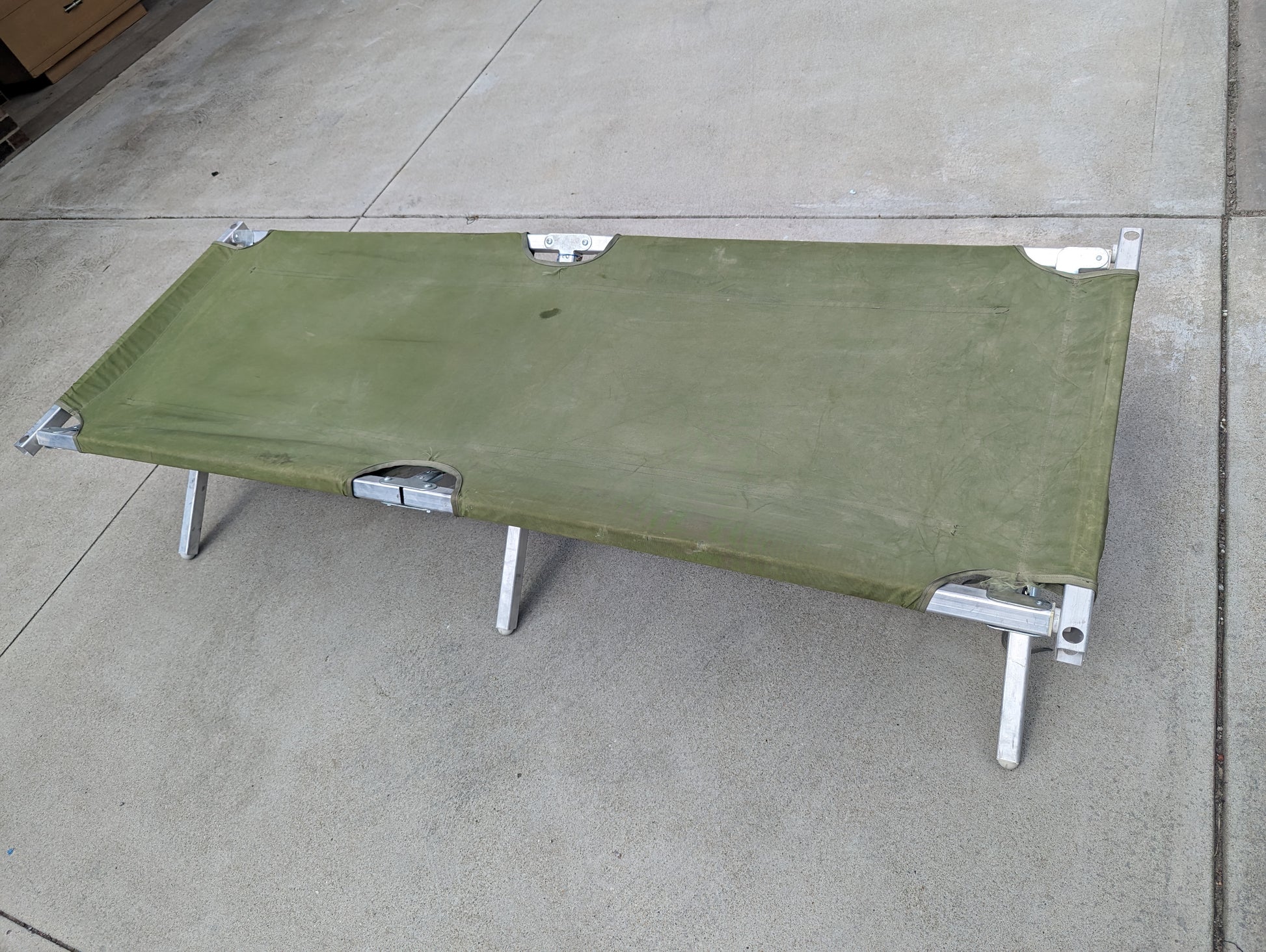A U.S. Military Surplus Folding Cot set up on a concrete floor, featuring a dark olive green fabric sleeping surface and a sturdy silver aluminum frame and legs.