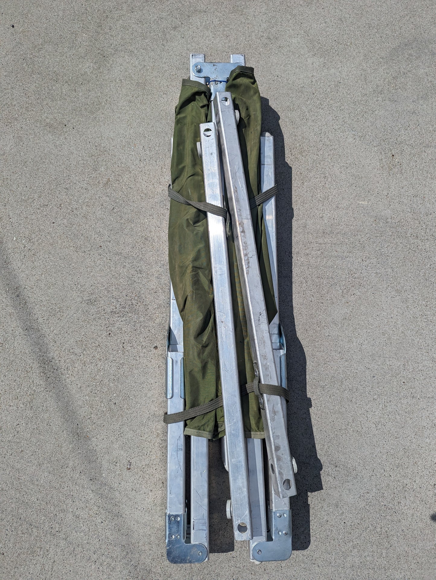 A U.S. Military Surplus Folding Cot in its collapsed state, featuring a dark olive green fabric surface wrapped around the silver aluminum frame components, resting on a gray concrete surface.