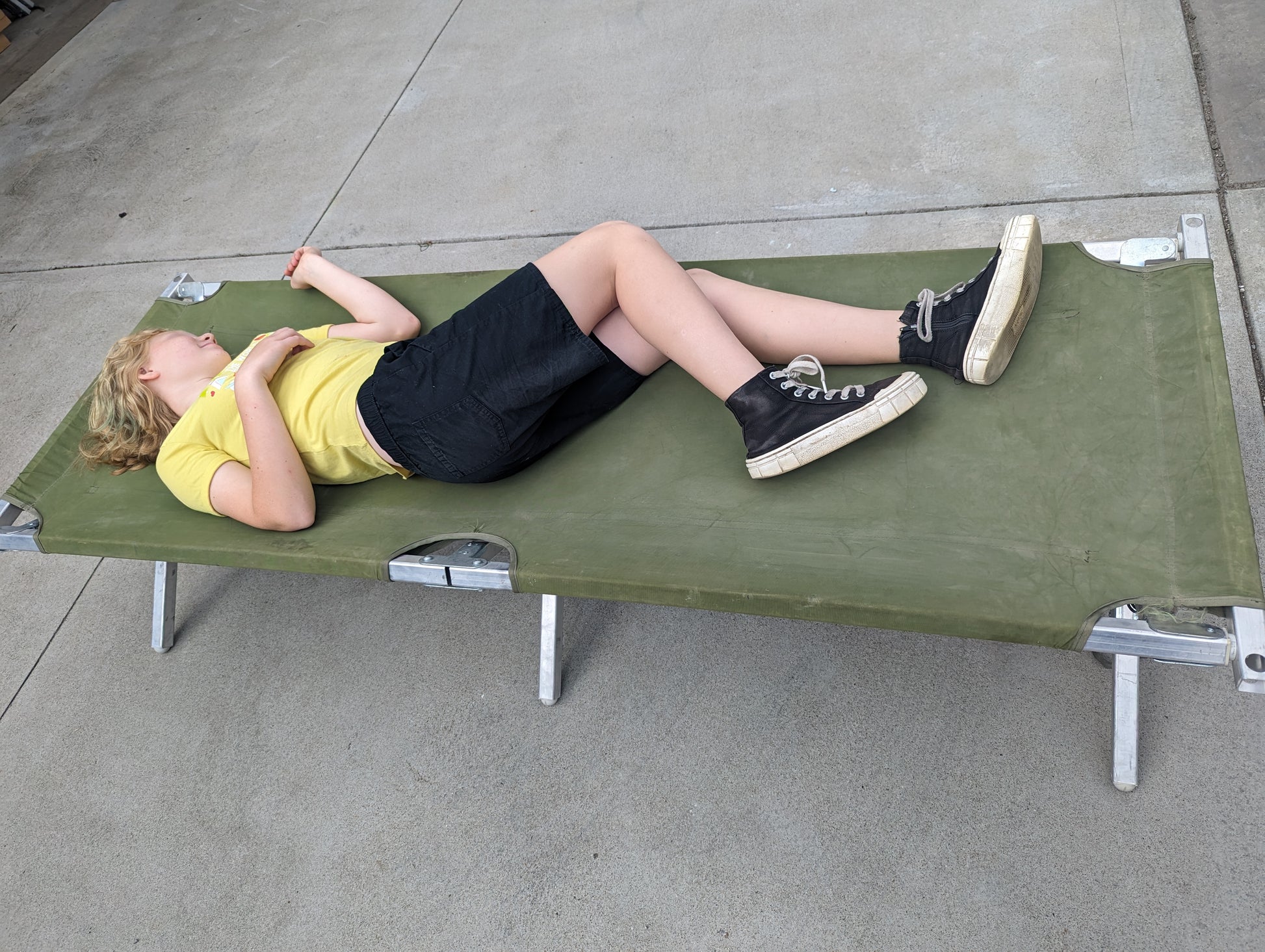 A person is lying on a U.S. Military Surplus Folding Cot with an olive green fabric surface and silver aluminum frame, demonstrating the cot's size and in-use capacity on a concrete surface.
