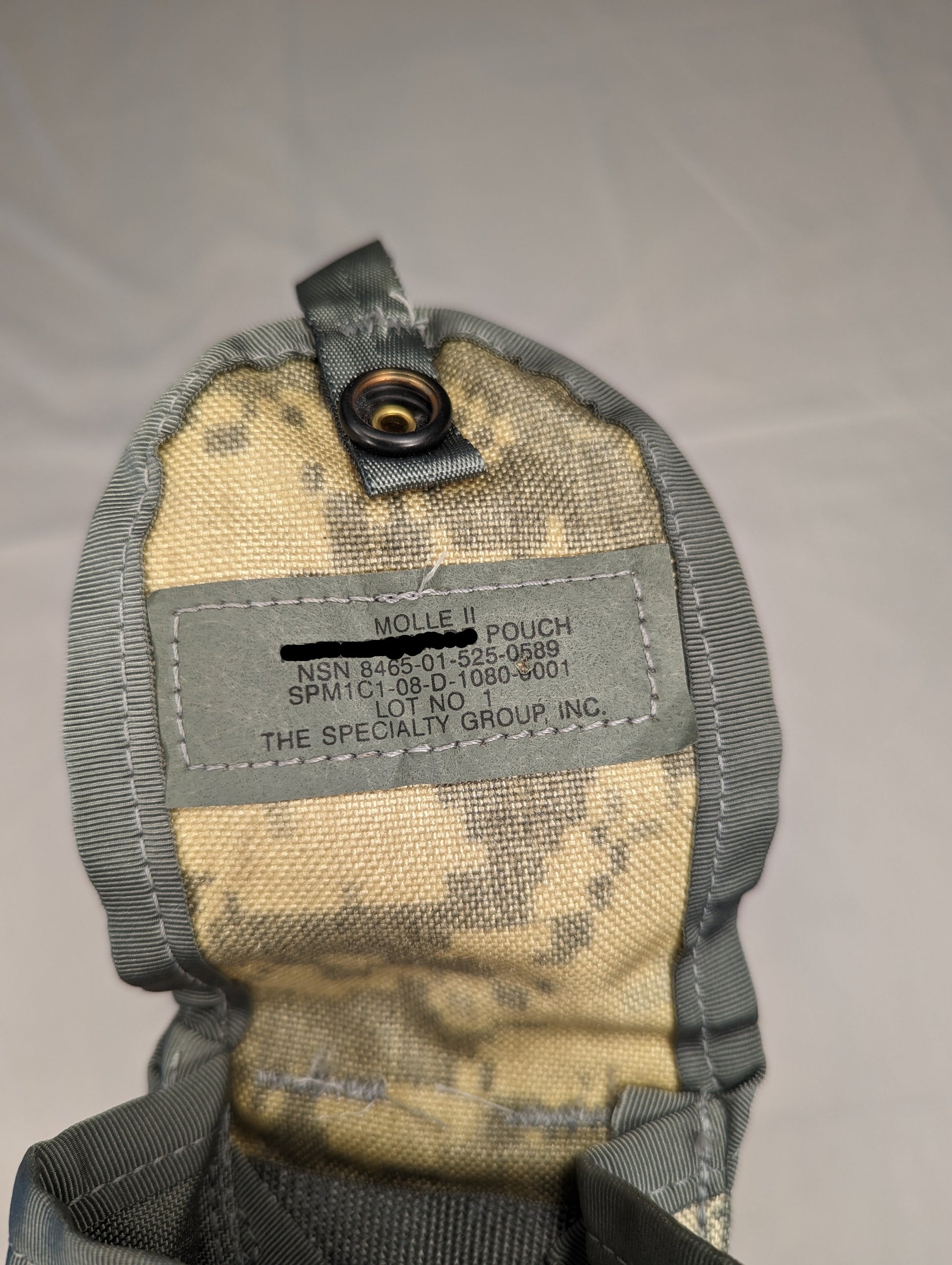 A close-up view of the open flap of a U.S. Military Surplus Hand Grenade Pouch in ACU digital camouflage, displaying the stitched gray issue tag with 'MOLLE II', NSN markings, and manufacturer 'The Specialty Group Inc.'.