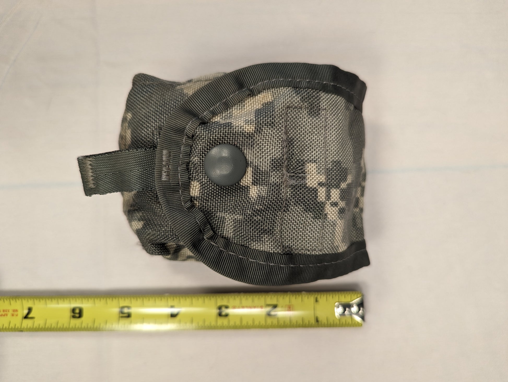 A top-down view of a single U.S. Military Surplus Hand Grenade Pouch in ACU digital camouflage, laid flat with a yellow tape measure positioned below to indicate width, featuring a snap-button flap.