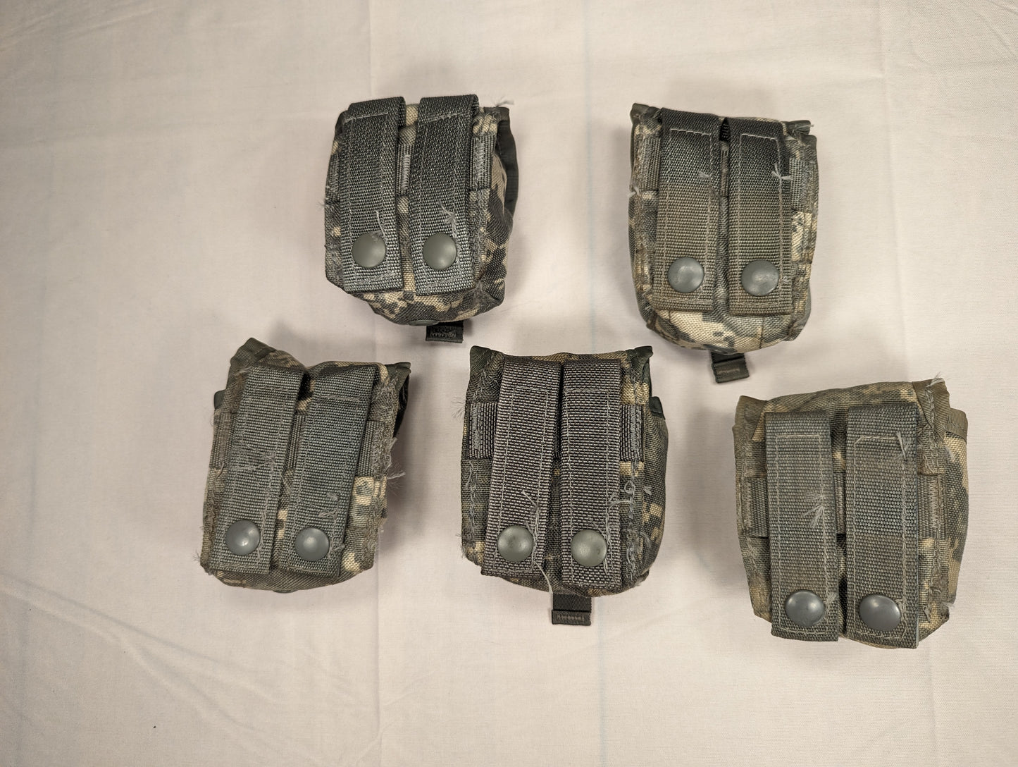 An overhead studio shot of five U.S. Military Surplus Hand Grenade Pouches in ACU digital camouflage, arranged in two rows on a white fabric background, each featuring a flap secured by two metal snap buttons.
