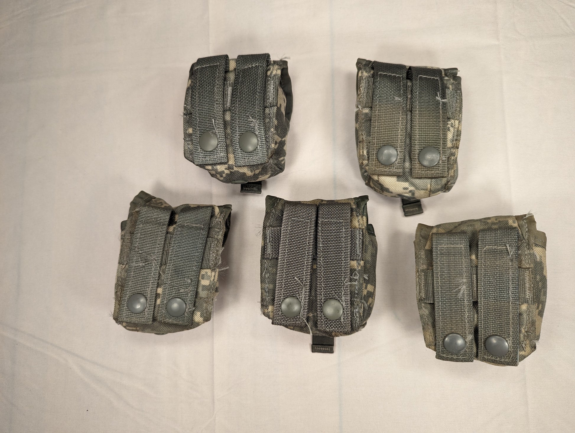 An overhead studio shot of five U.S. Military Surplus Hand Grenade Pouches in ACU digital camouflage, arranged in two rows on a white fabric background, each featuring a flap secured by two metal snap buttons.