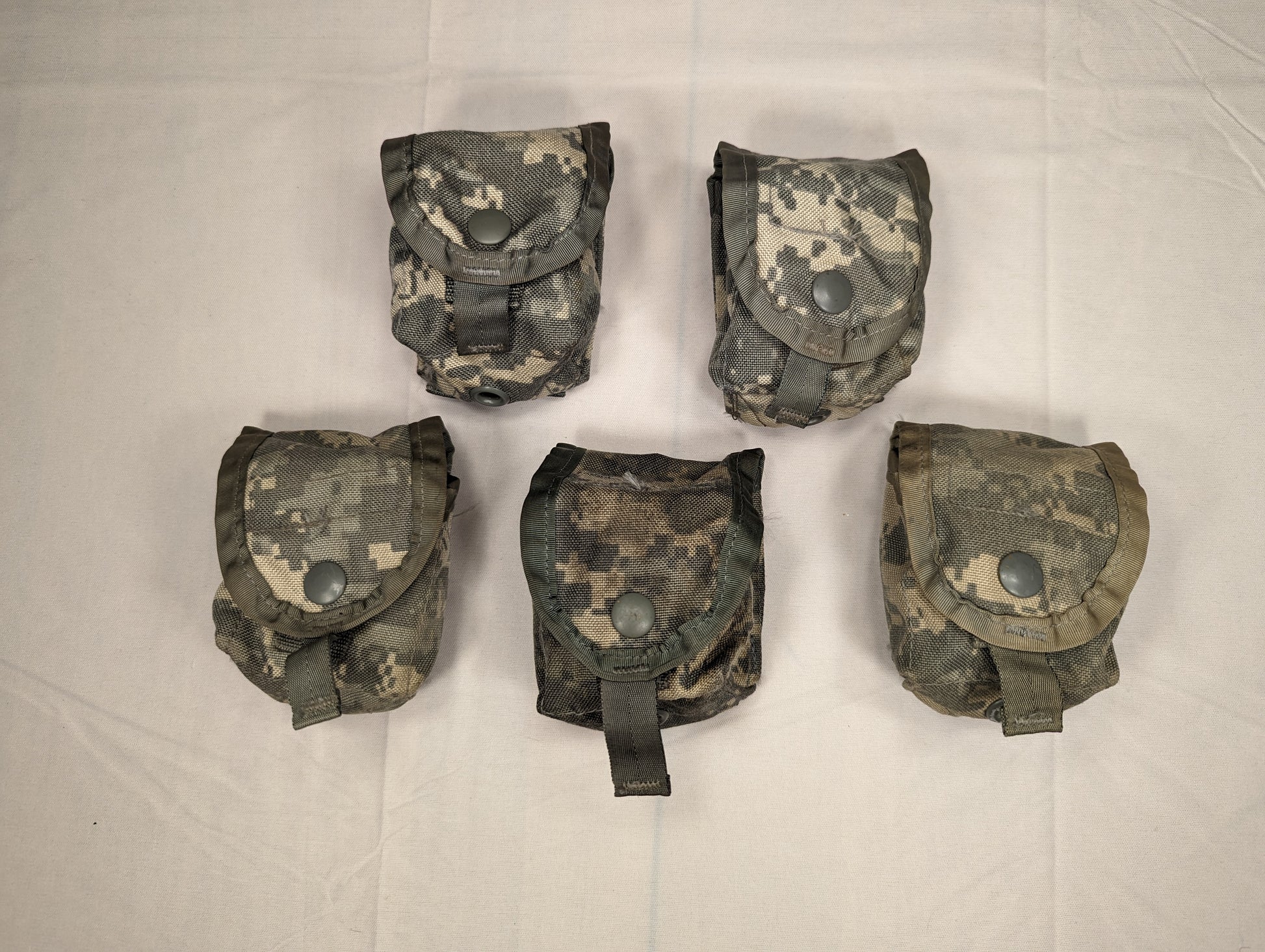 An overhead studio shot of five U.S. Military Surplus Hand Grenade Pouches in ACU digital camouflage, arranged in two rows on a white fabric background, each featuring a snap-button flap.