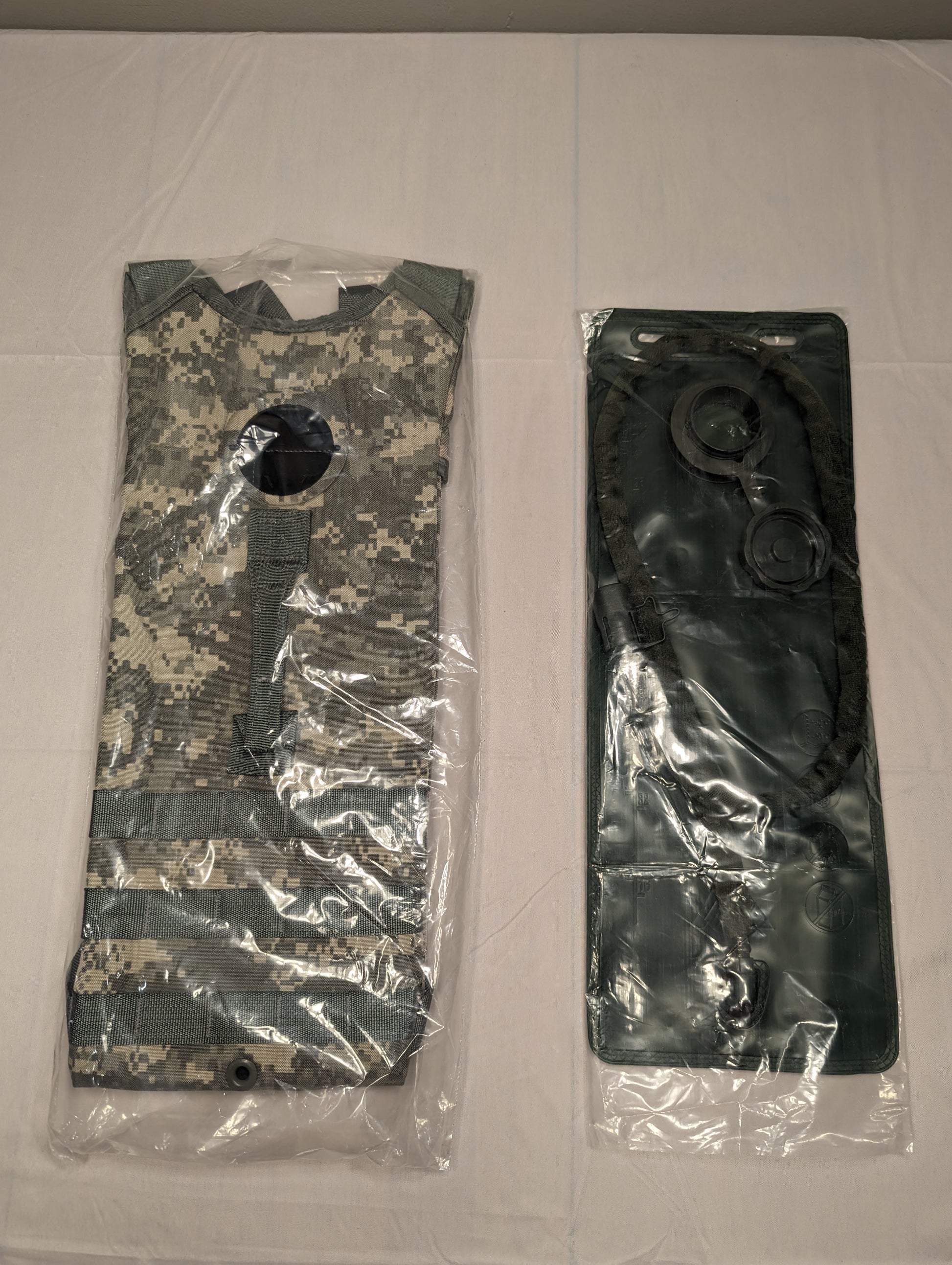 Two U.S. Military Surplus Hydration System components, including the ACU digital camouflage carrier shell and the black 100oz bladder, sealed separately in clear plastic bags and displayed side-by-side.