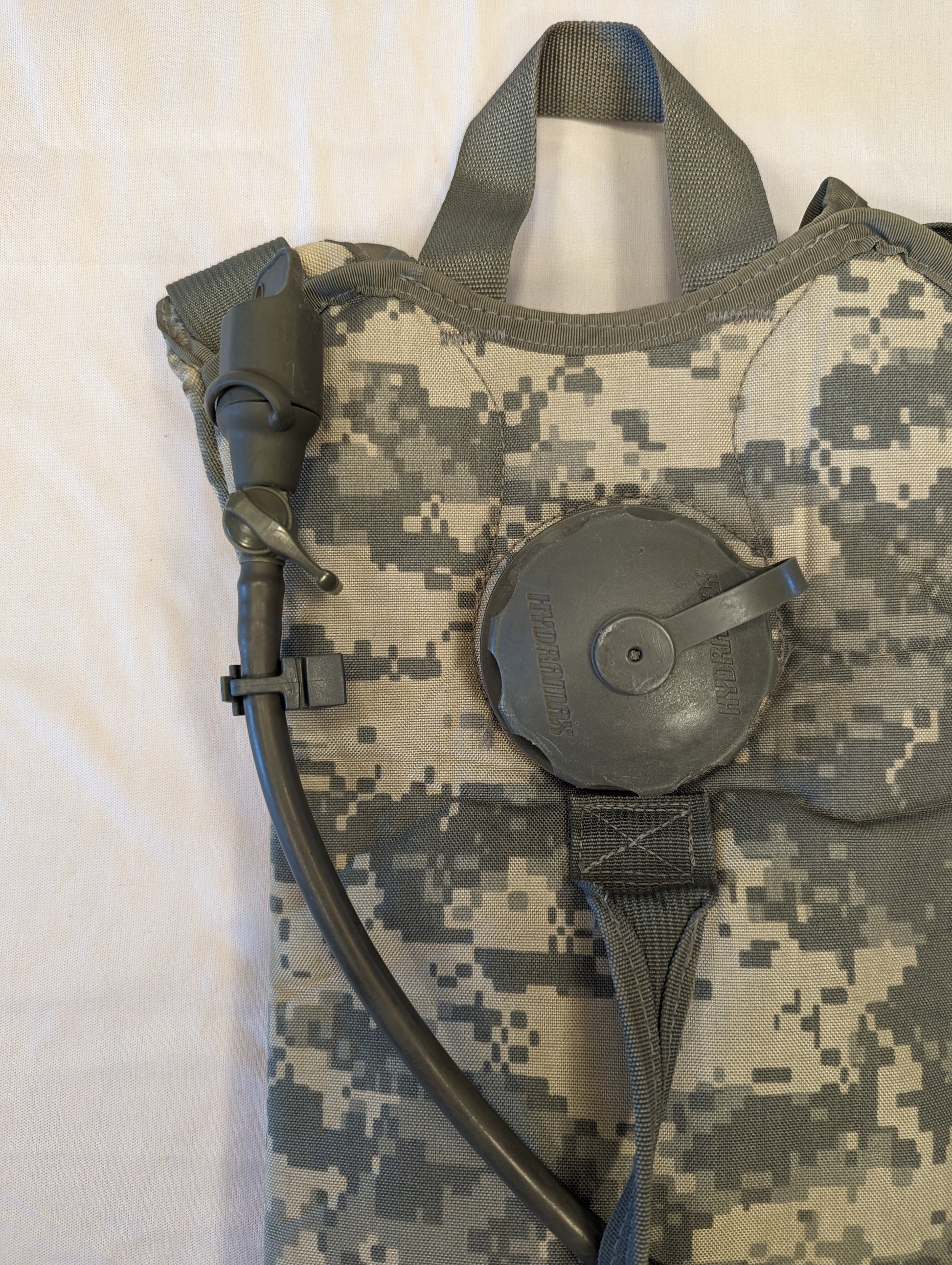 A close-up view of the U.S. Military Hydration Carrier in ACU digital camouflage, highlighting the large black filler cap, the attached drinking tube, and the carrying handle.